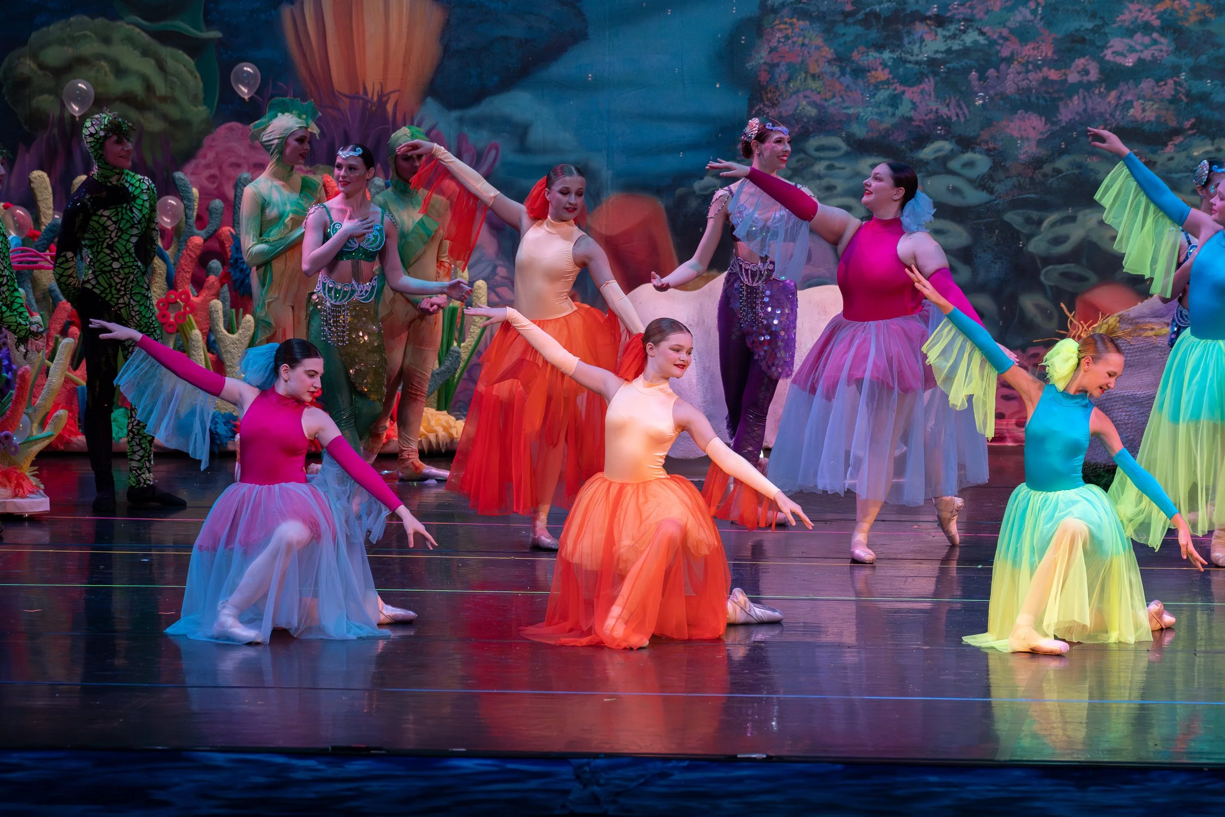 Young dancers in colorful costumes perform a ballet on stage with an ocean-themed backdrop.