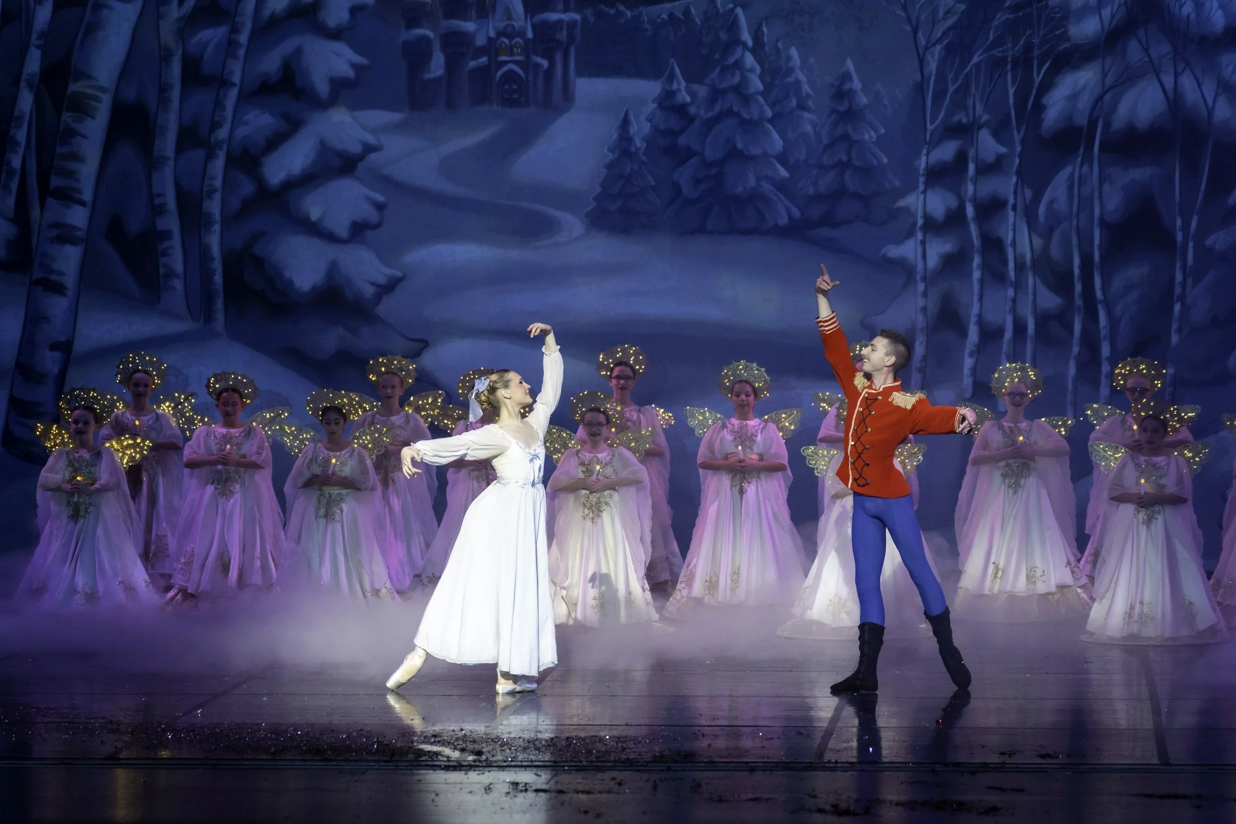 A ballet performance with dancers dressed as characters from a holiday scene, including a female dancer in a white dress and male dancer in a red coat, with a backdrop depicting a snowy winter landscape and a group of ballerinas dressed as angels wit