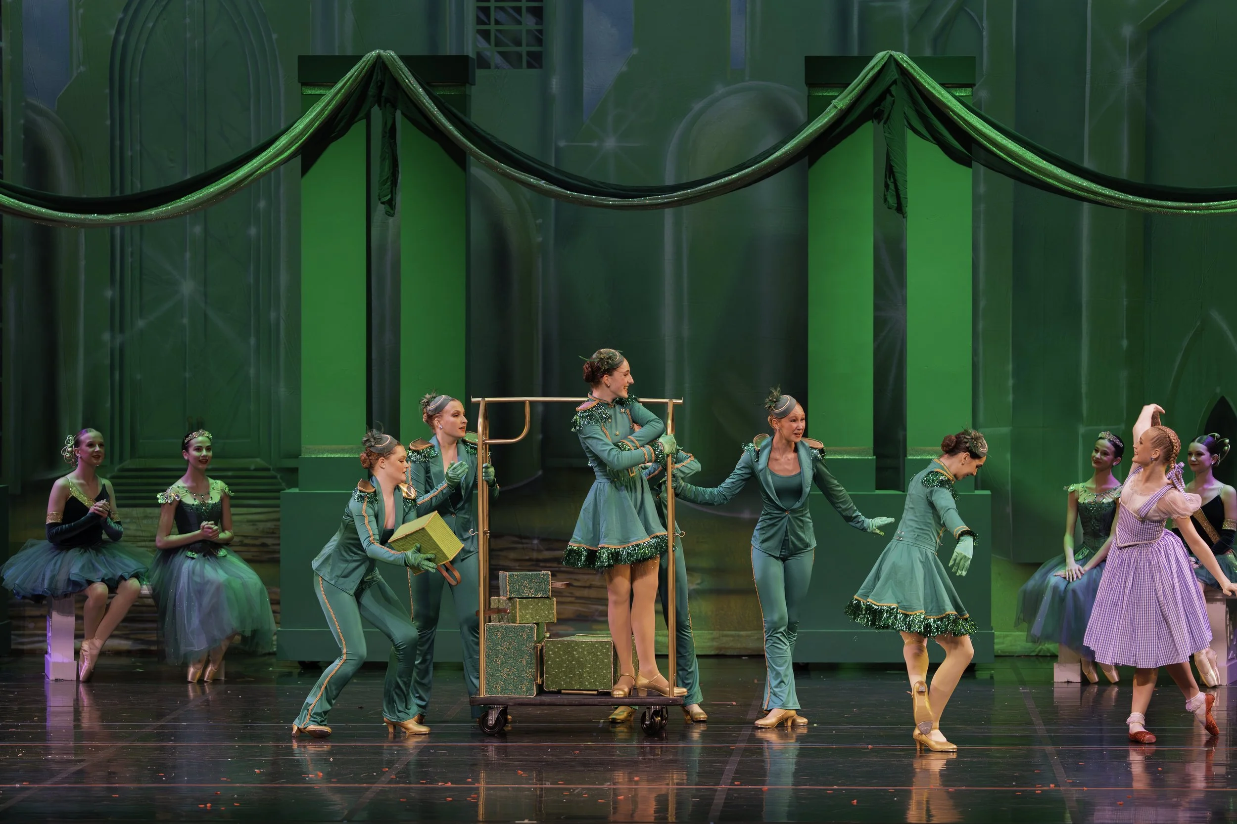 Ballet dancers performing on stage with a green backdrop, some on pointe and others surrounding a cart with a woman in a teal dress standing on it.
