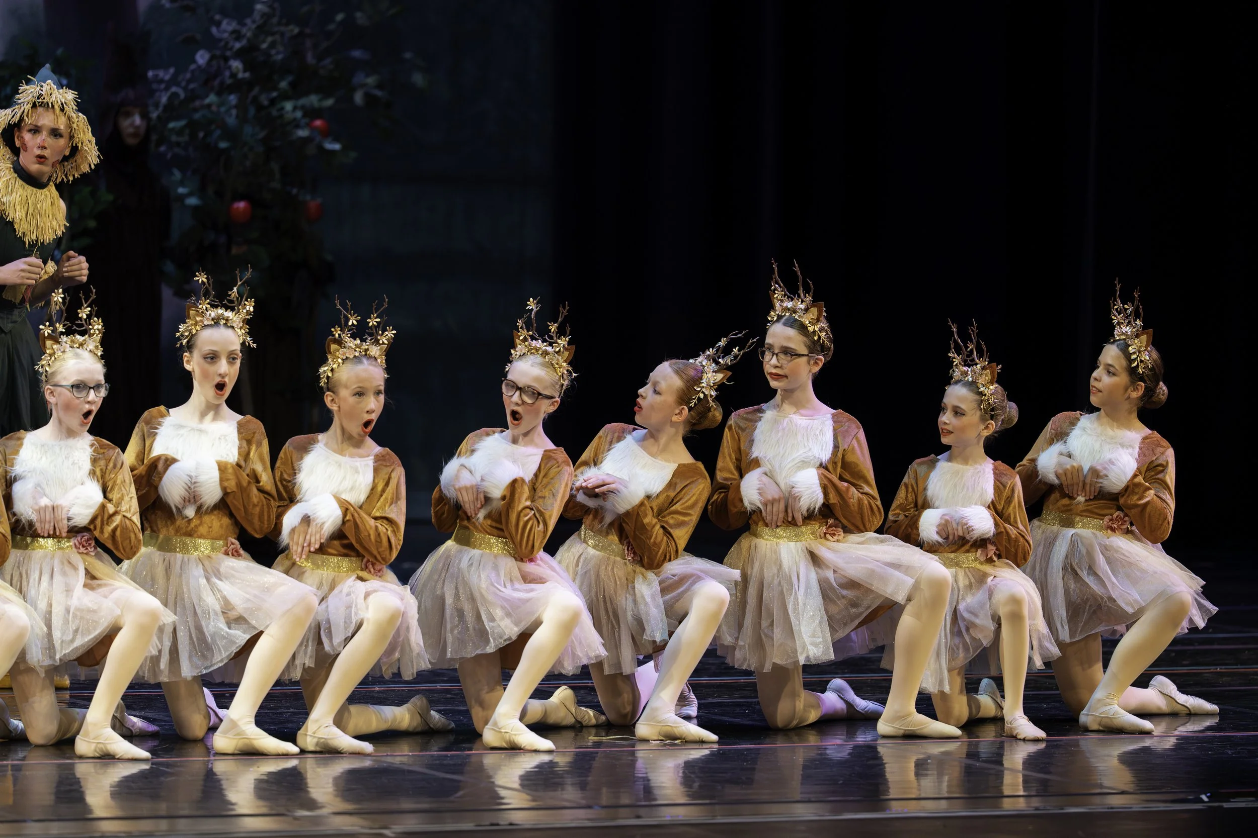 Young girls dressed as reindeer performing a ballet on stage, with some girls kneeling and others standing, wearing costumes with antler headpieces and plush white chest accents.