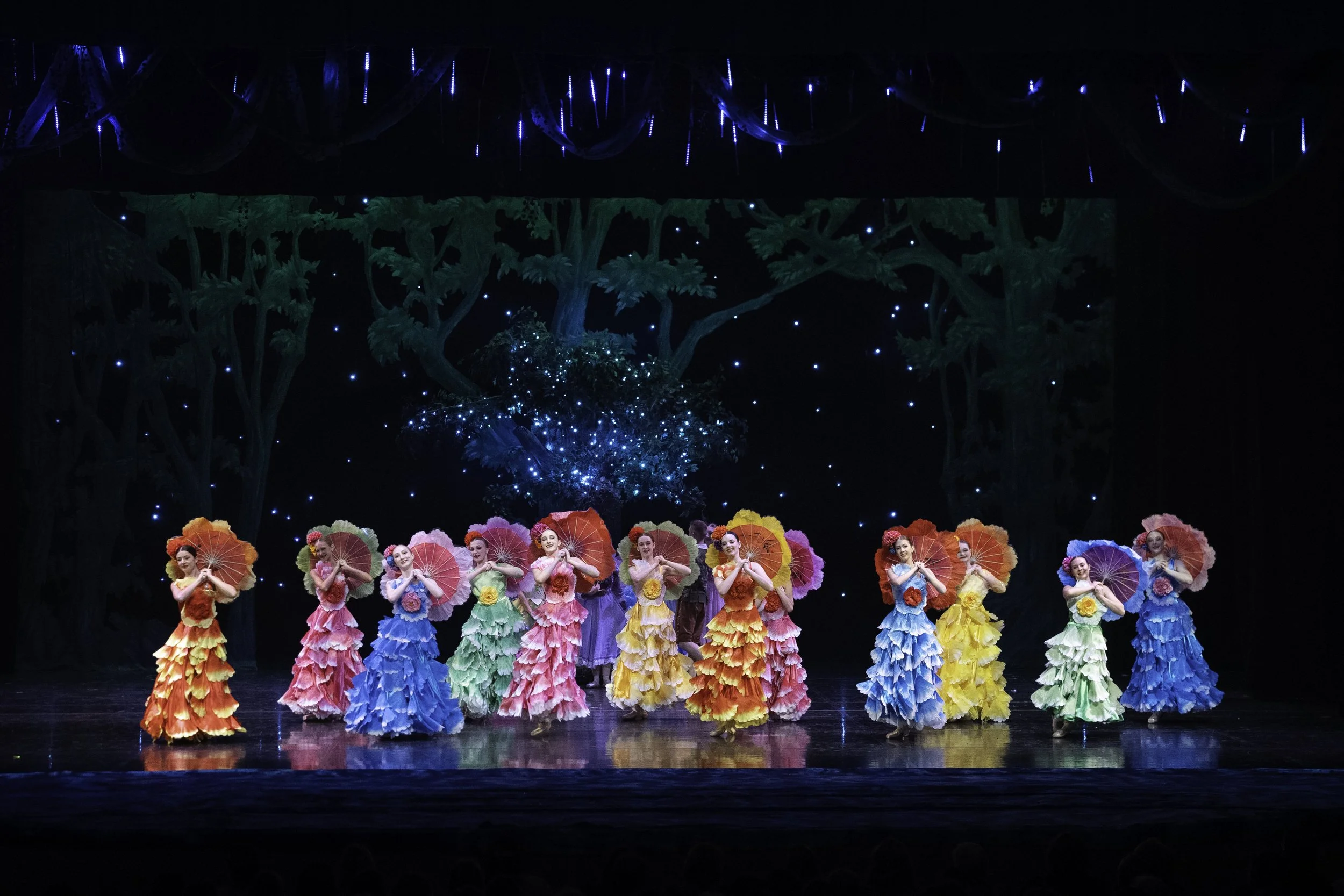 Stage with ten women in colorful ruffled dresses holding parasols, performing a dance under a starry backdrop with forest-like scenery.