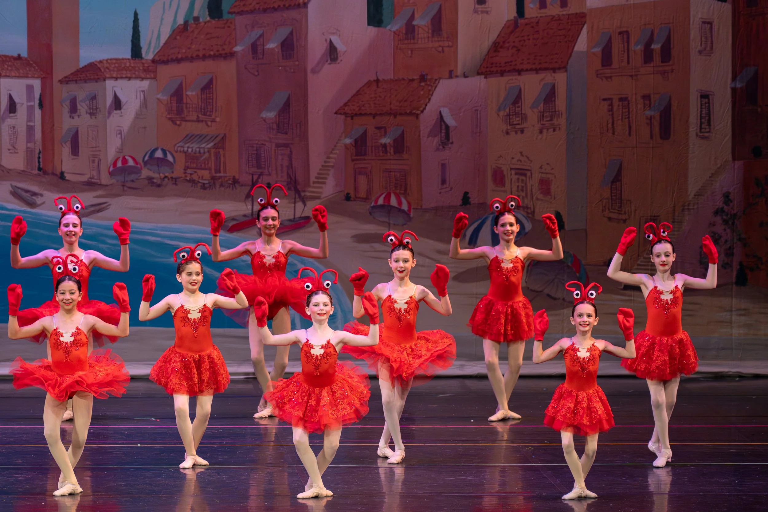 A group of young ballet dancers in red costumes with ladybug-themed headbands practicing on stage with a painted backdrop of a seaside town.
