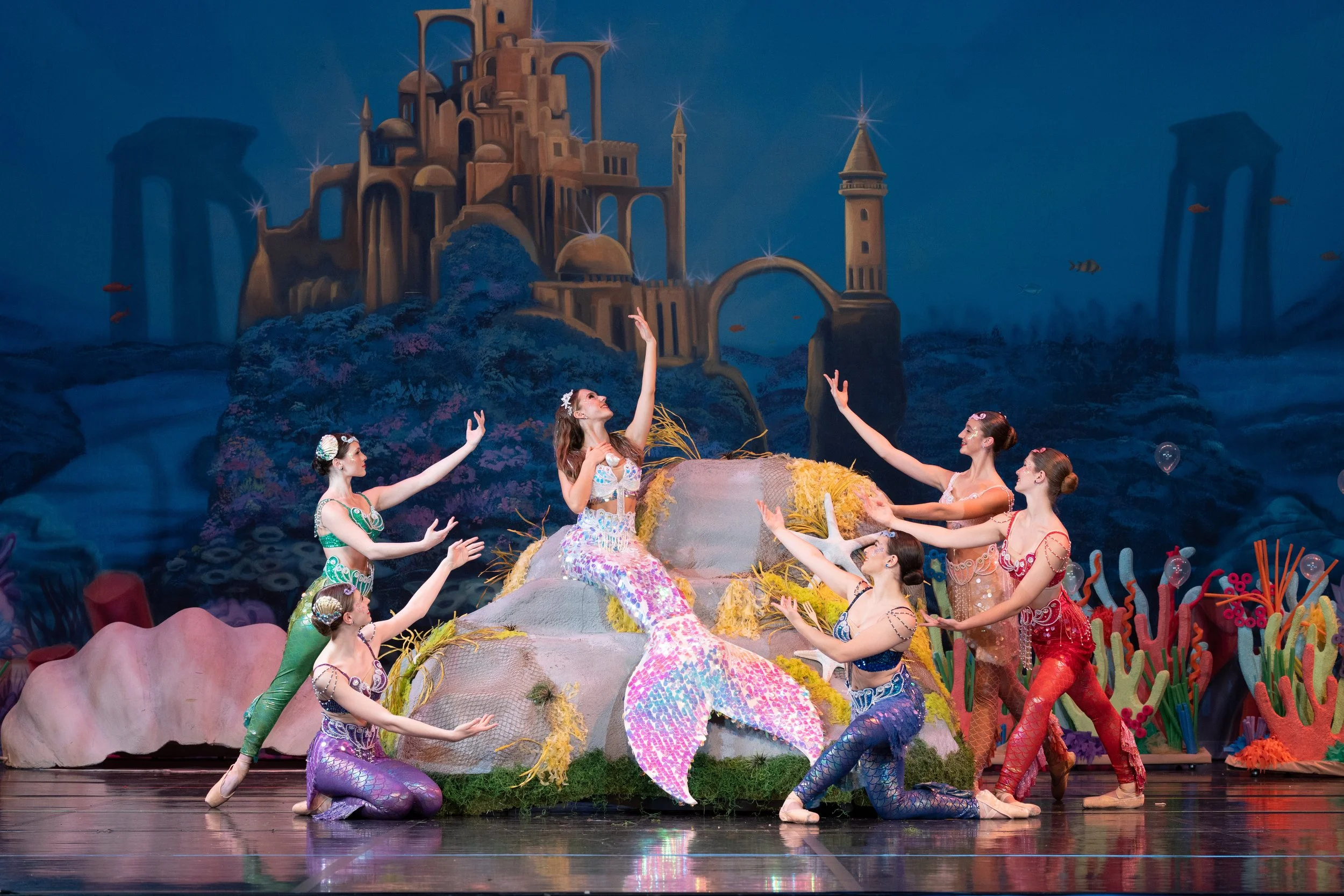 Ballet dancers dressed as mermaids performing on stage with a colorful underwater-themed backdrop featuring a castle, coral, and fish.