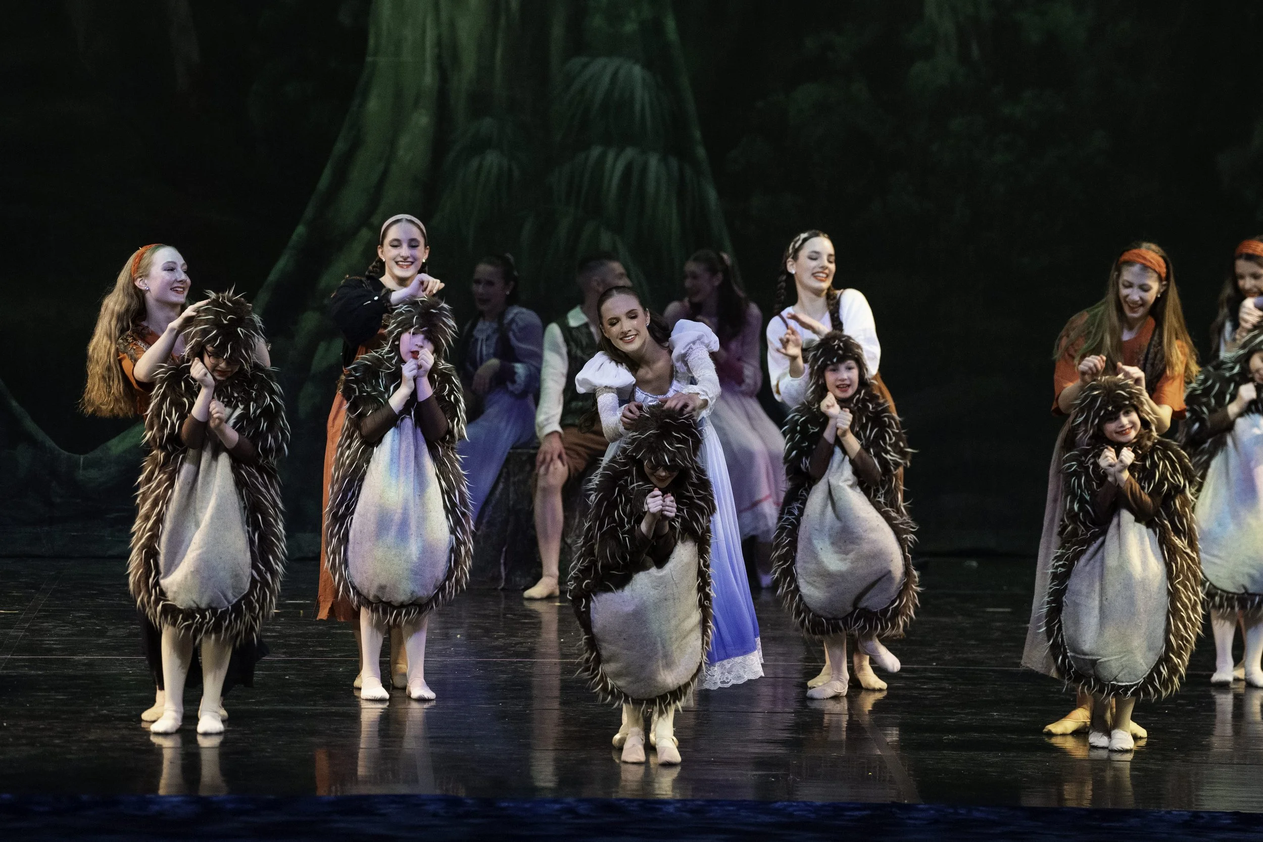Children in costumes performing on stage, dressed as animals with fluffy costumes and white paws, in a theatrical production set with a backdrop resembling a forest.