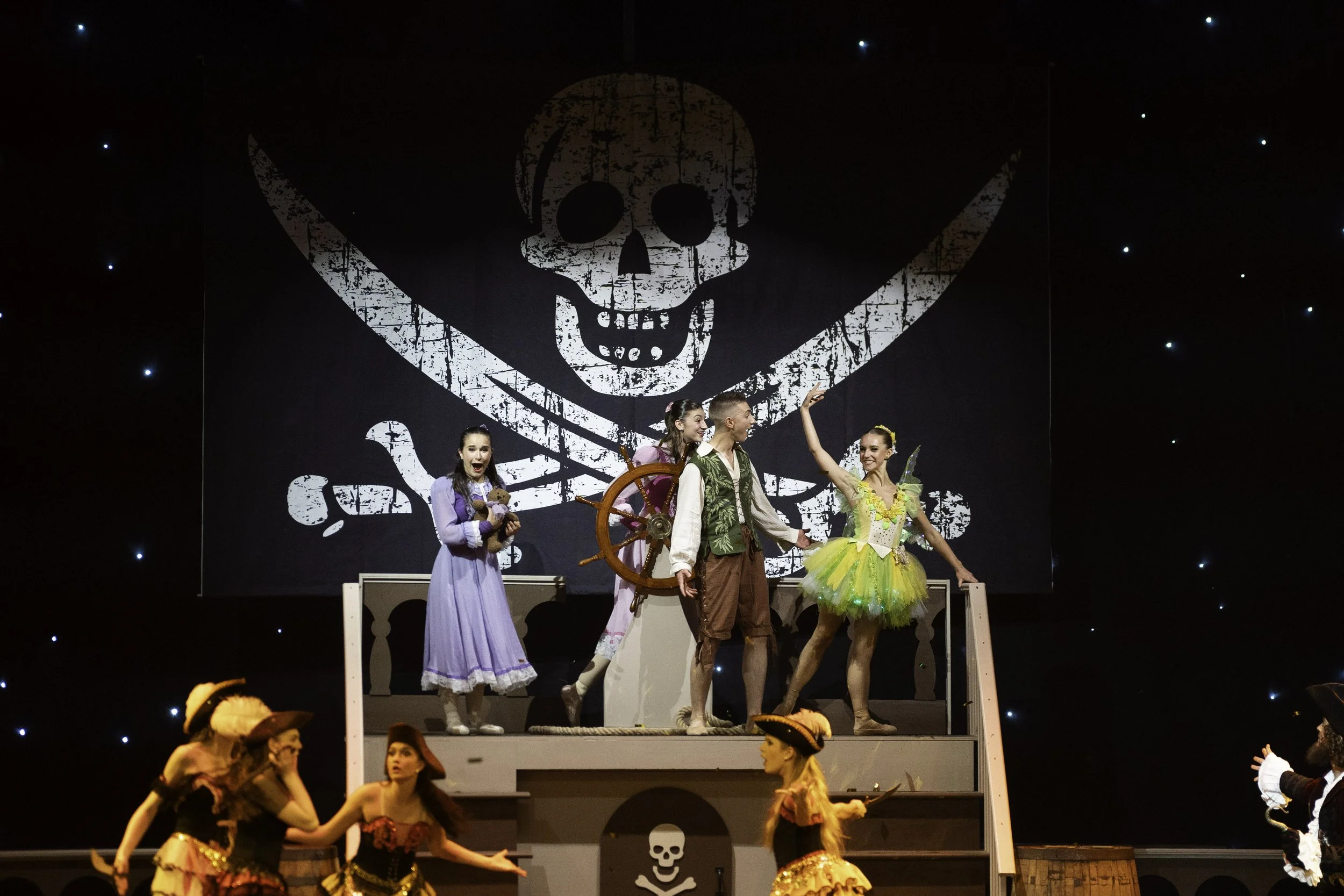 Children performing on stage in a play with a large skull and crossbones backdrop, dressed in costumes representing pirates and fairy tale characters.