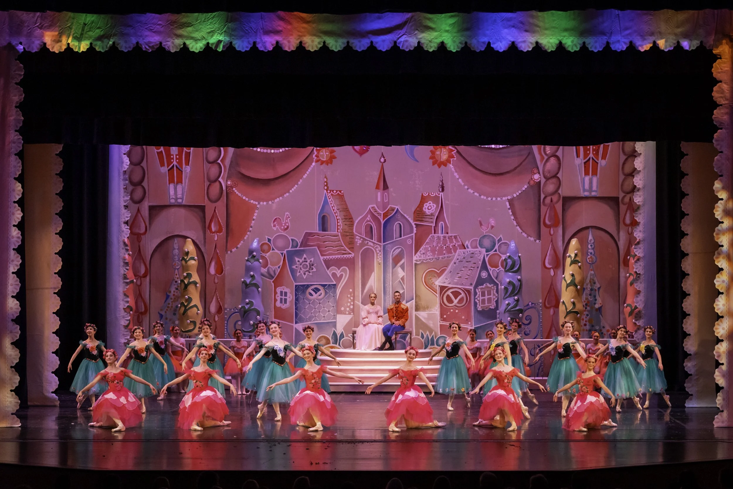 A stage performance featuring ballet dancers in pink and green costumes with a colorful, whimsical castle backdrop and elaborate set design.