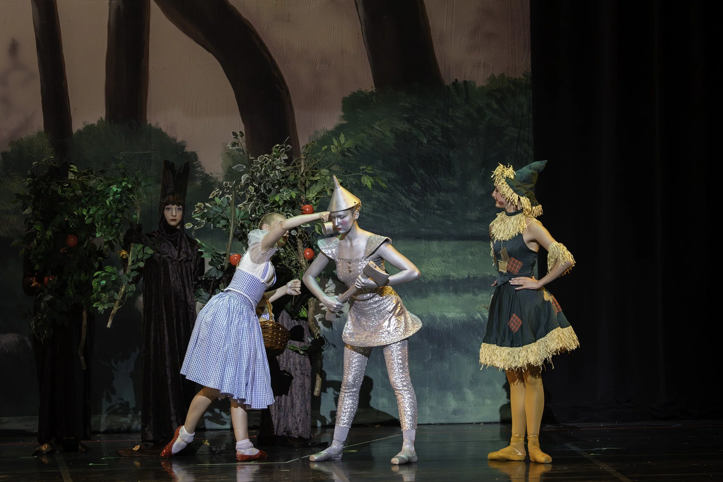 Children's theater performance of The Wizard of Oz. A girl dressed as Dorothy is placing a silver cone hat on a clown-like figure dressed as the Wizard. A woman dressed as Glenda the Good Witch stands to the right observing, with a backdrop of painte