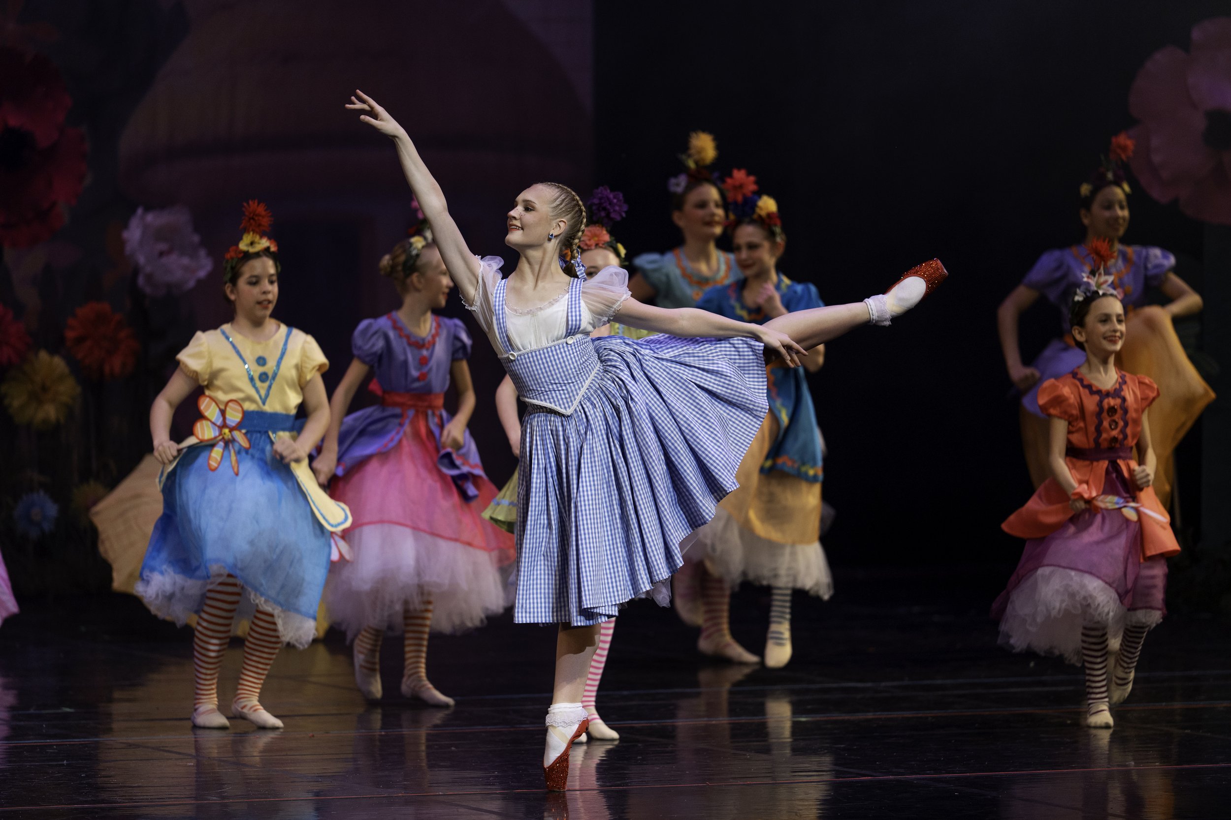 Young ballerinas in colorful costumes performing on stage with floral decorations in the background.