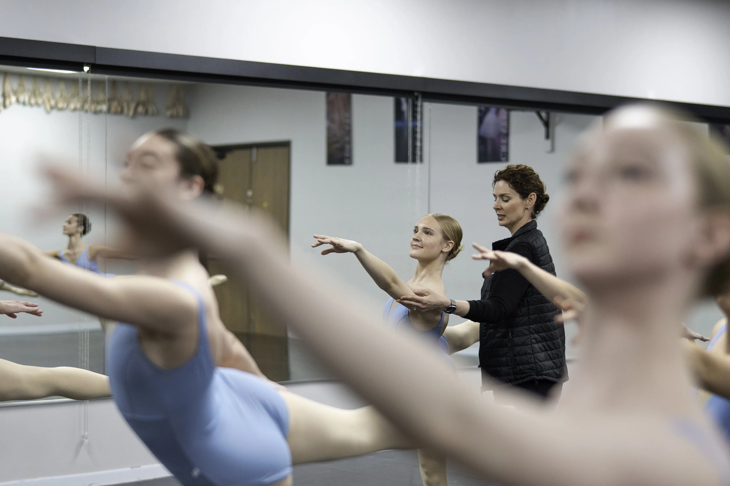 Ballet instructor helping young dancers stretch during a class in a dance studio with mirrors.