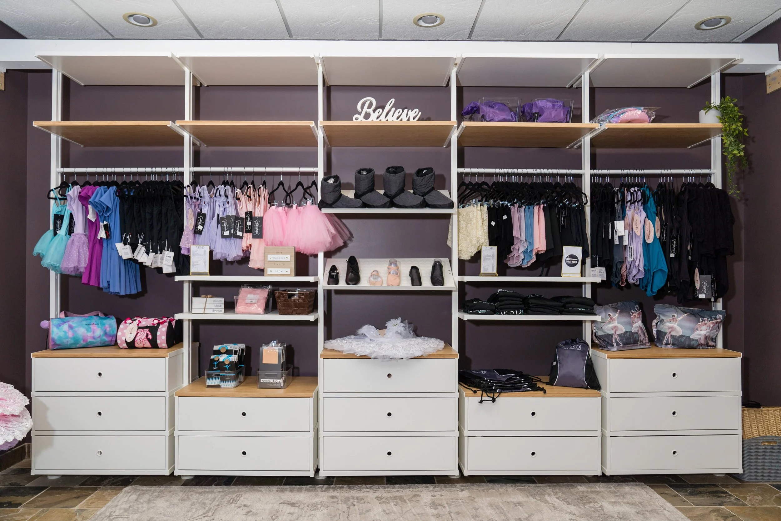 Display shelf with ballet dancewear and accessories, including tutus, leotards, shoes, and bags, against a purple wall with decorative 