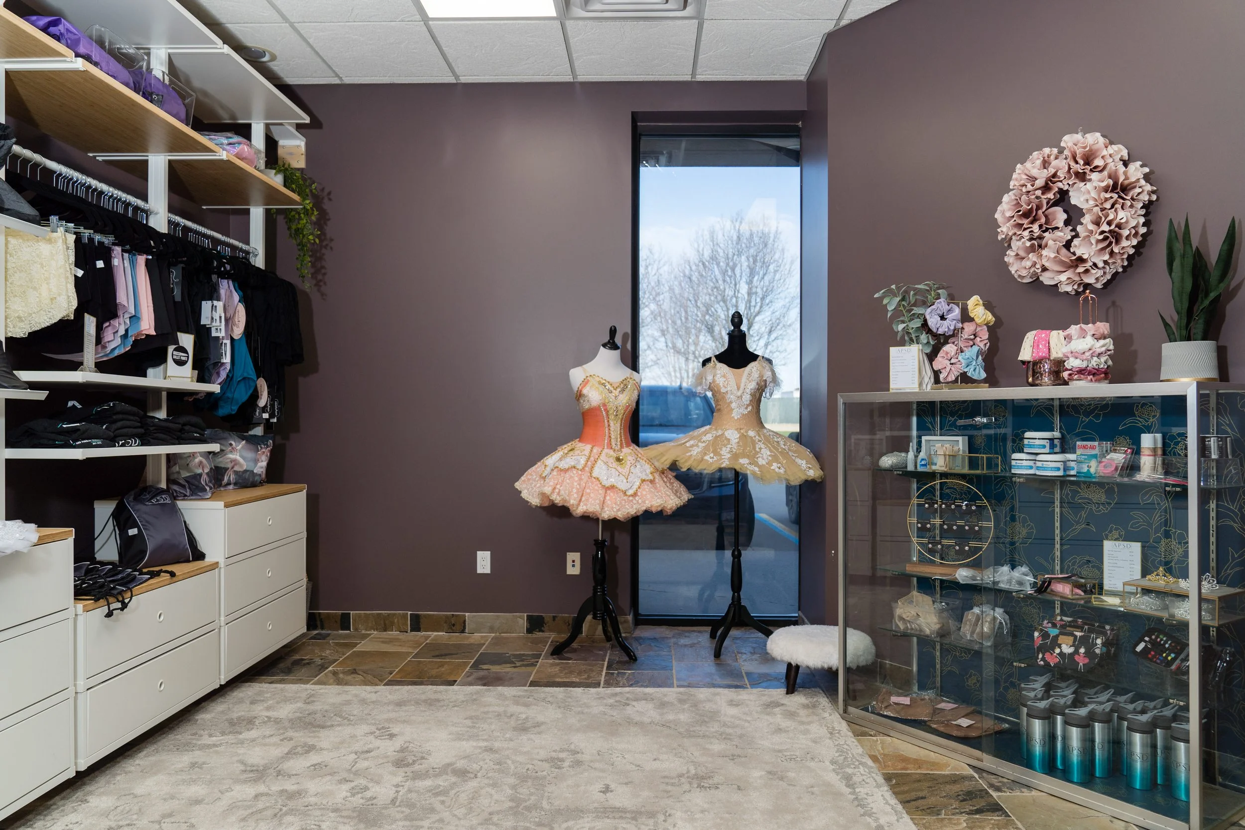 Inside a store, two ballet costumes on mannequins near a window, with shelves of accessories and decor, purple walls, and a glass display case.