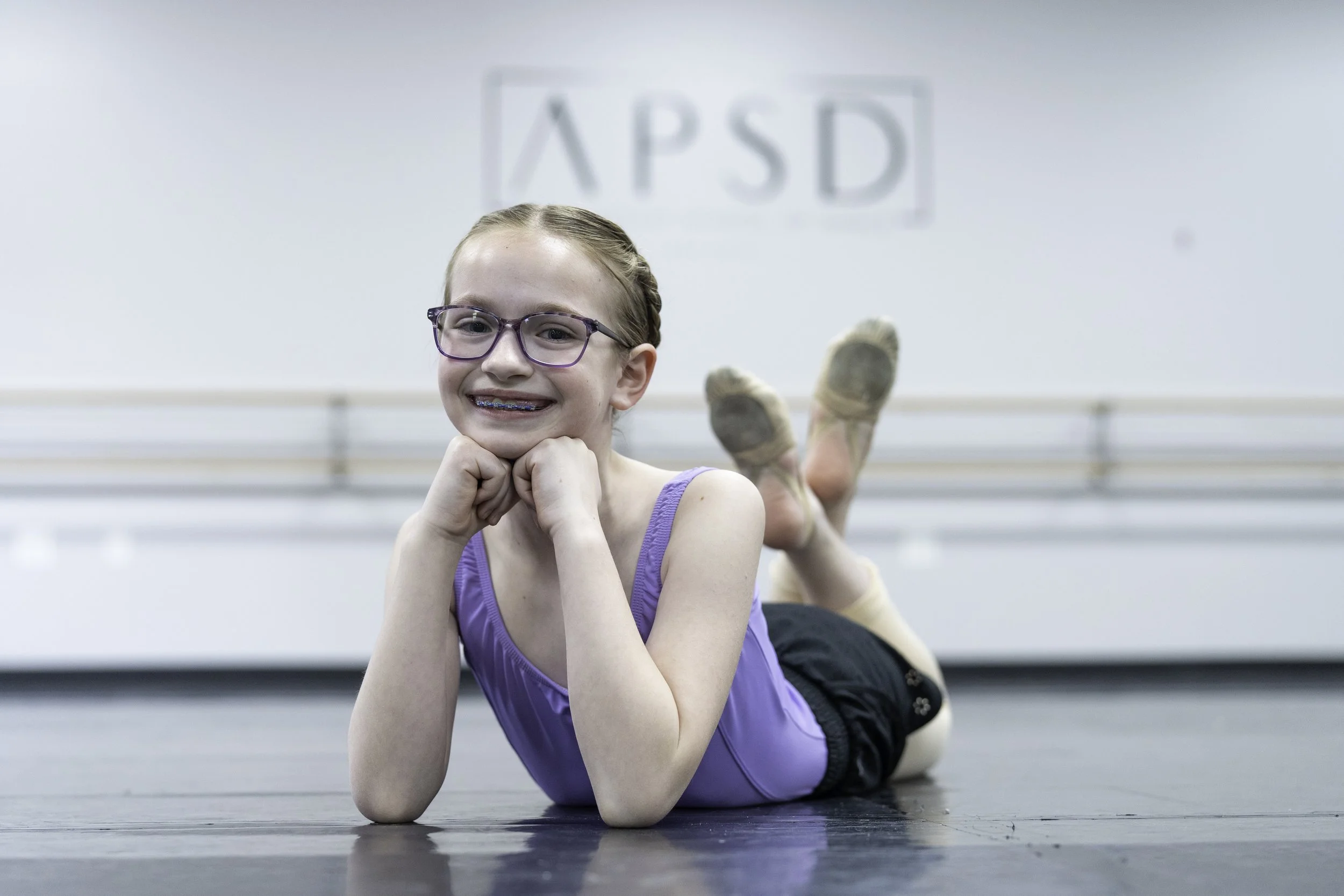 Young girl in a purple tank top and glasses lying on a dance studio floor, smiling, with ballet barres and a PSD sign in the background.