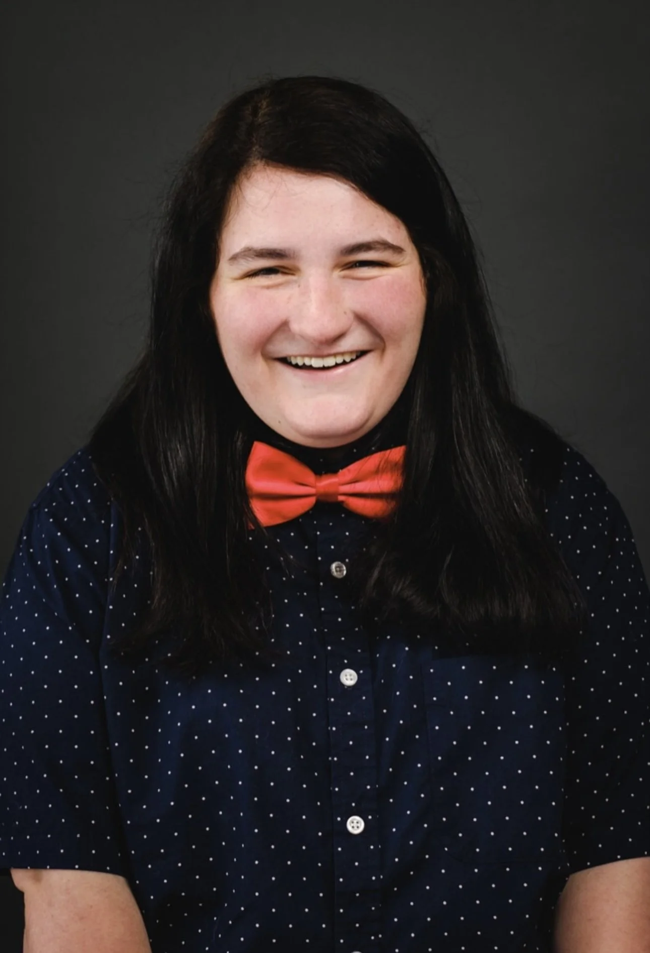 Portrait of a young woman with long dark hair, smiling, wearing a red bow tie and a navy blue shirt with white polka dots, against a dark background.