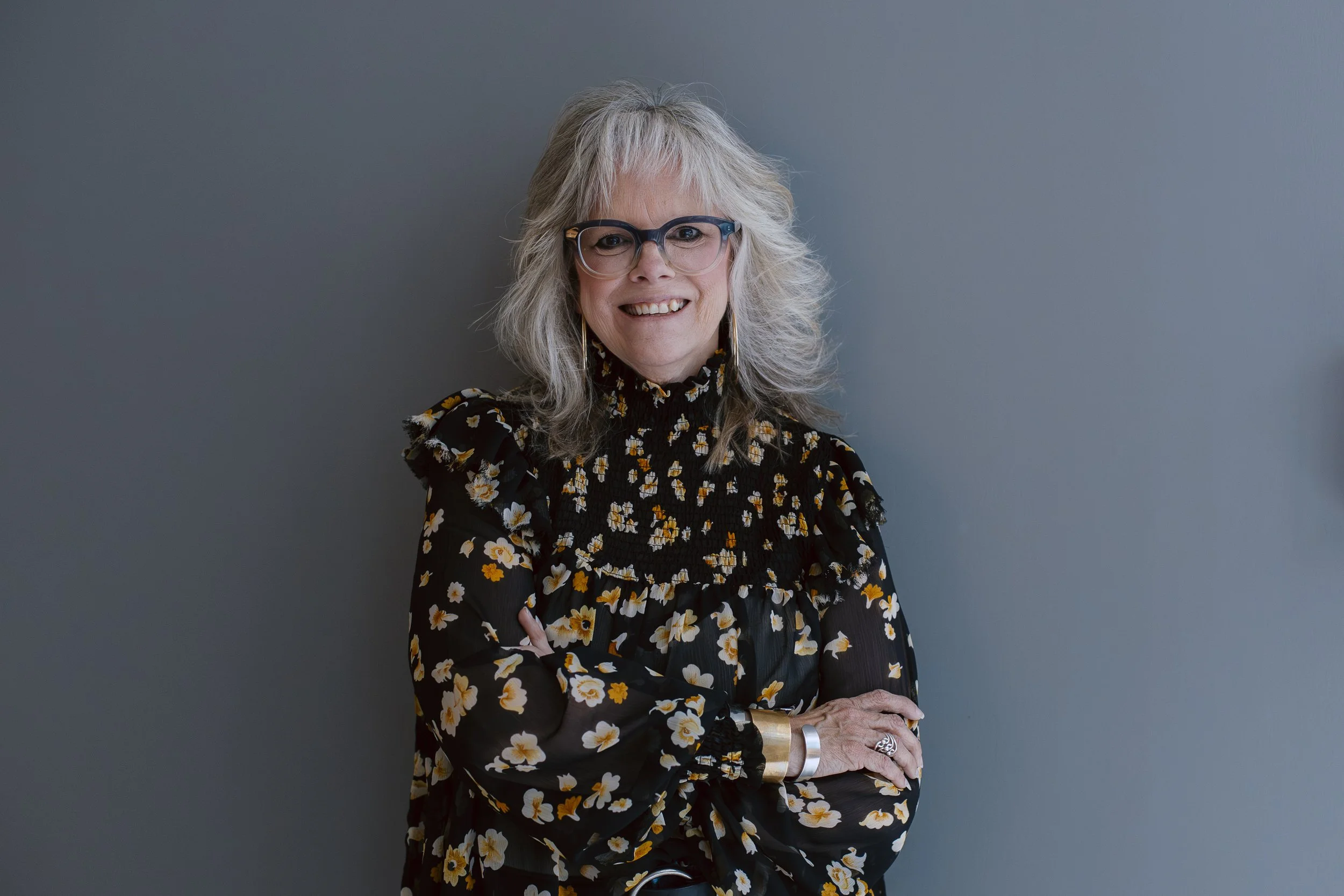 A woman with gray hair, glasses, and a floral patterned black blouse, smiling and standing with her arms crossed against a plain gray wall.