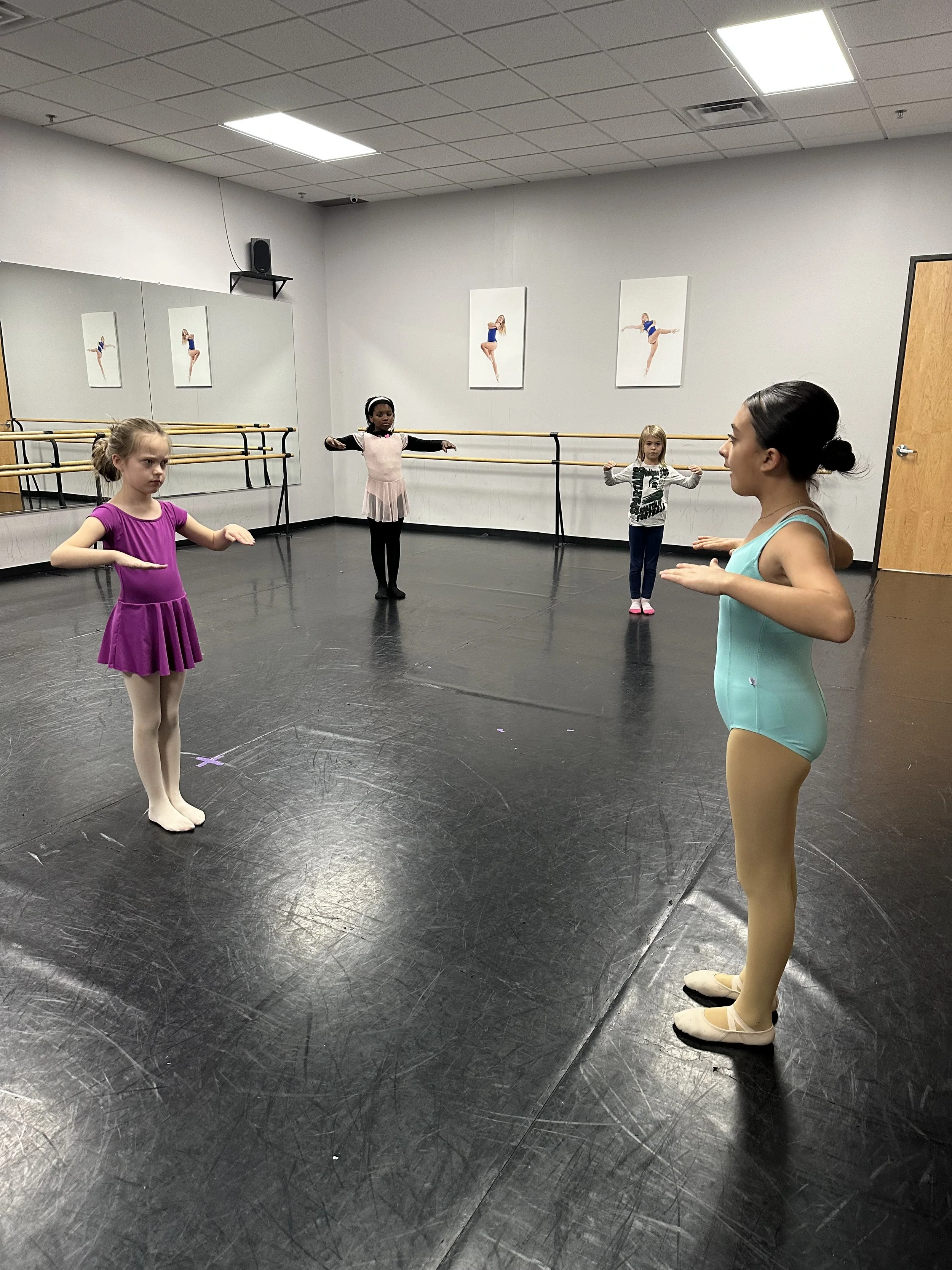 Young ballet students in a dance studio, practicing ballet positions with an instructor observing. The studio has ballet barres, framed dance photos, a mirror, and a black floor.