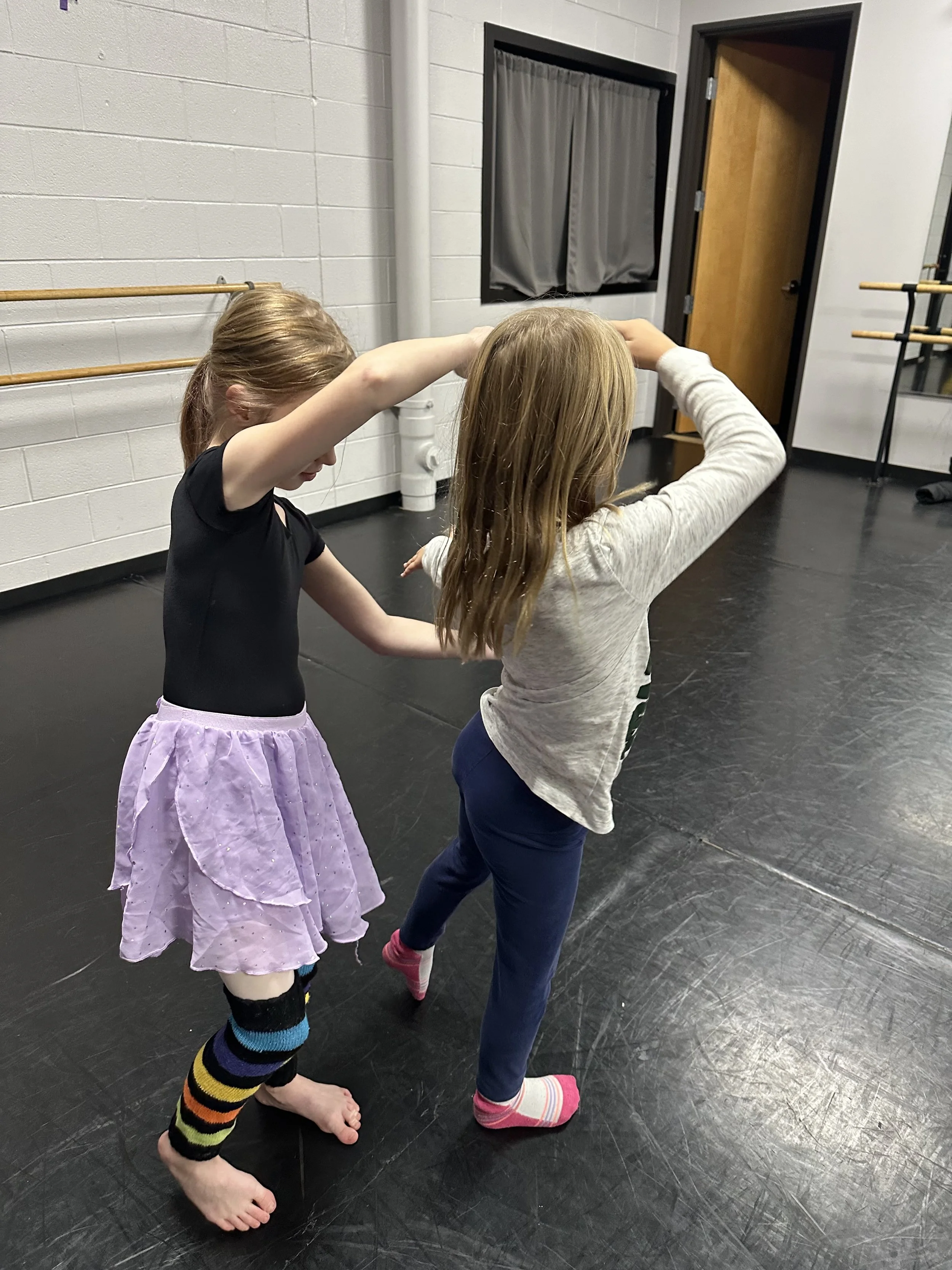 Two young girls practicing ballet or dance at a studio, with one girl helping the other stretch or warm up.