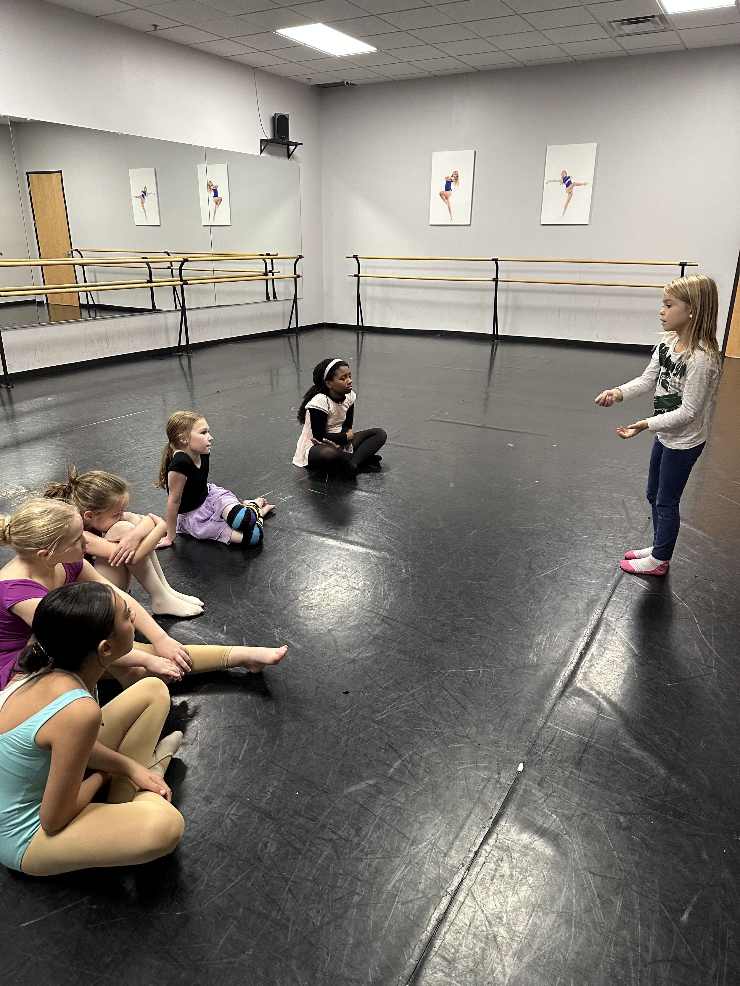 A ballet dance class with young girls sitting on the floor and a girl standing and speaking to them. The studio has ballet barres, a large mirror, and framed ballet photos on the wall.