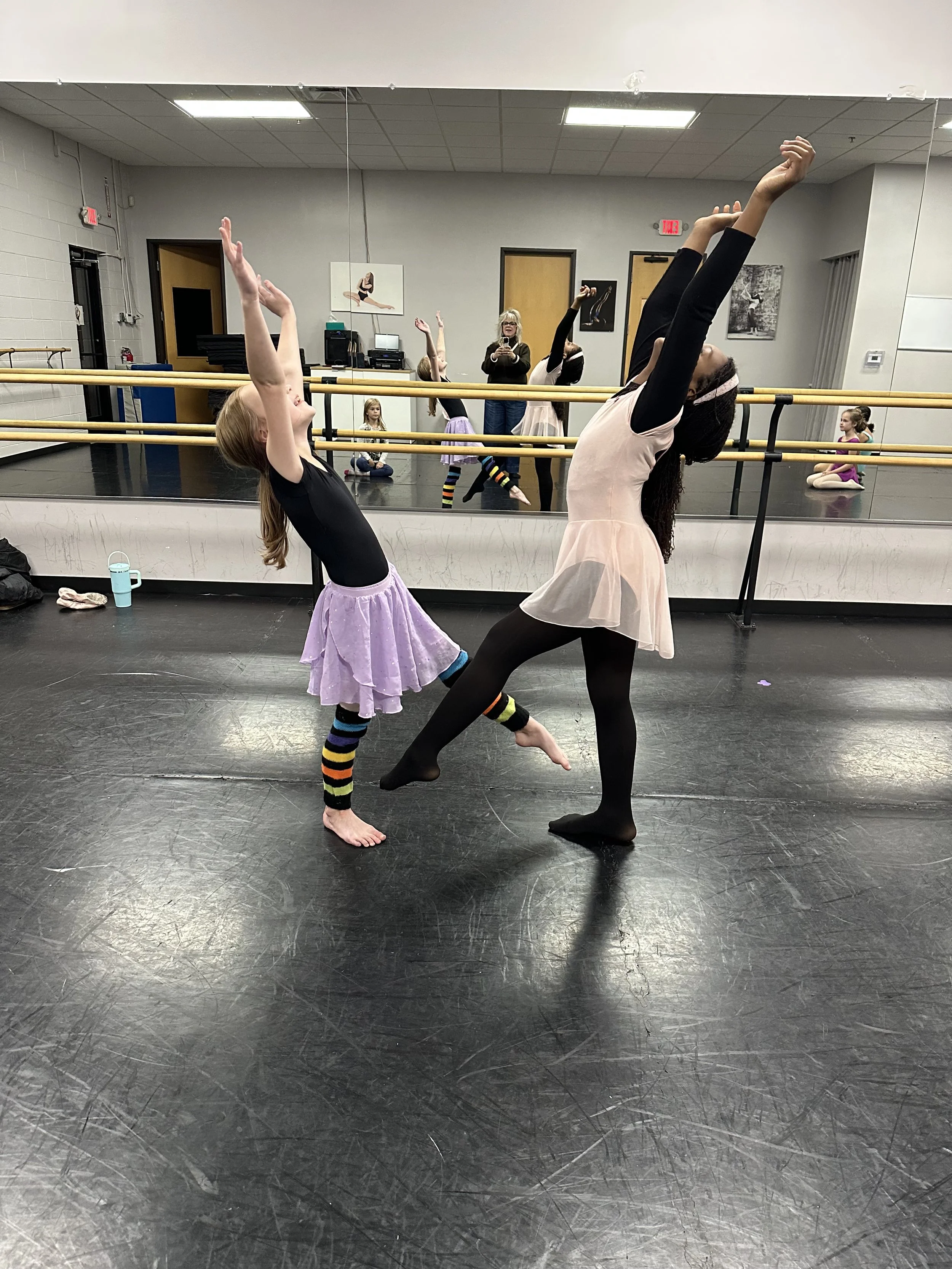 A ballet class in progress with a young girl and an instructor performing a stretch at the barre. The girl is wearing a purple skirt and striped leg warmers, while the instructor is dressed in black tights and a light pink leotard. Other young studen