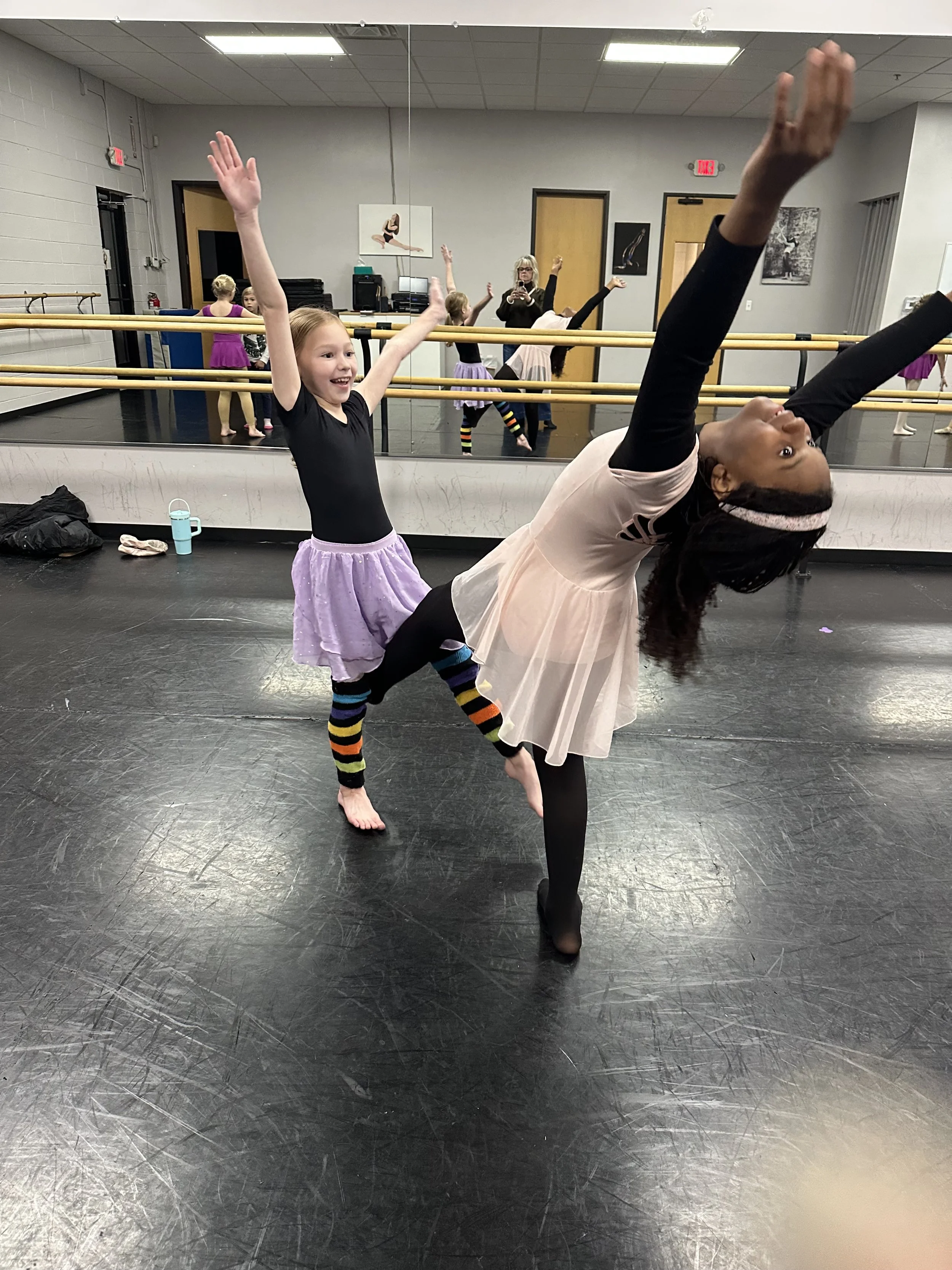 A young girl in a black shirt and purple tutu is smiling and raising her arms in a dance studio, assisted by an adult woman dressed in black and a light pink skirt, who is in a dance pose with her back arched and arms extended.