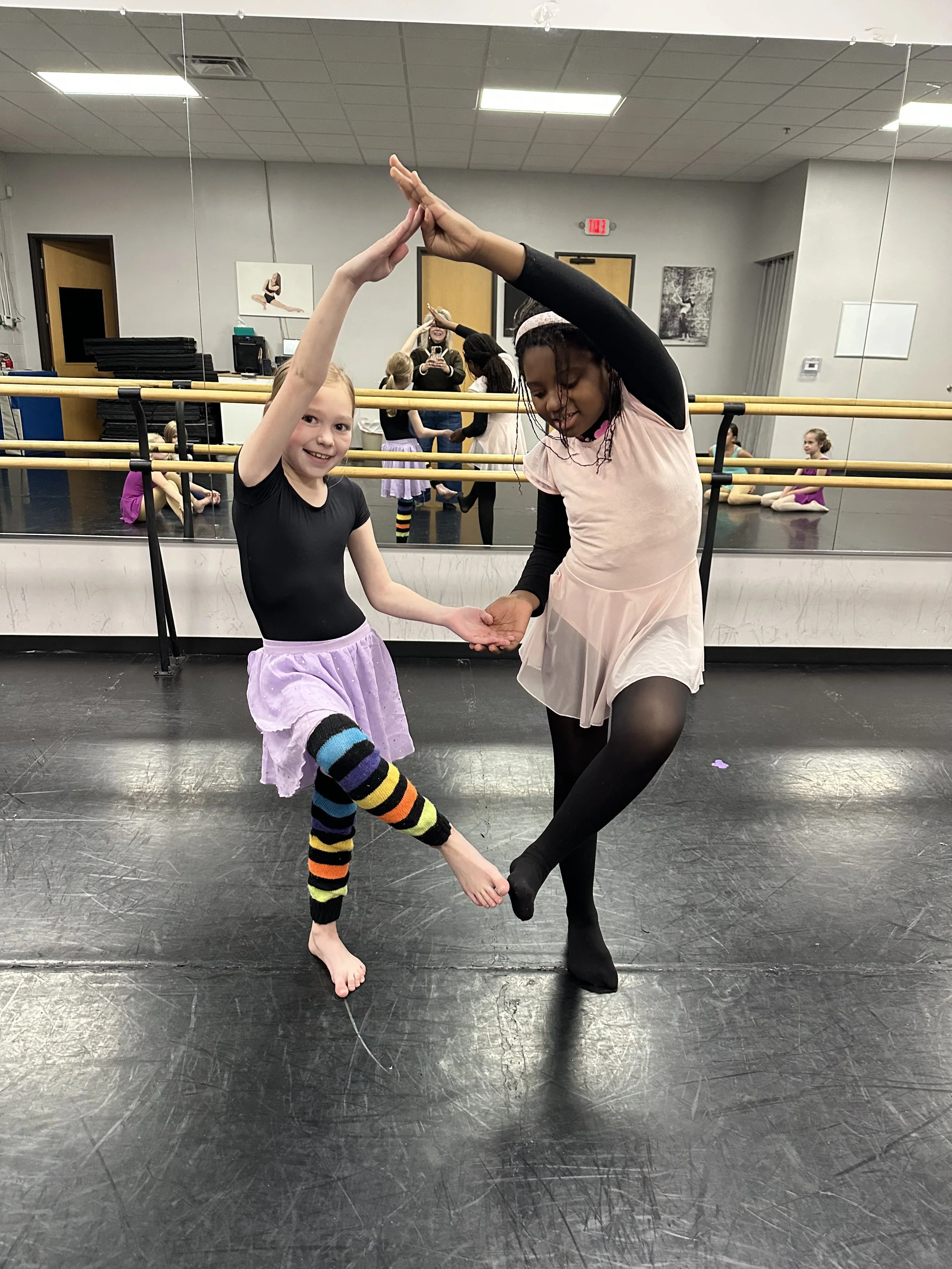 Two young girls practicing ballet in a dance studio, smiling and holding hands while balancing on one leg.