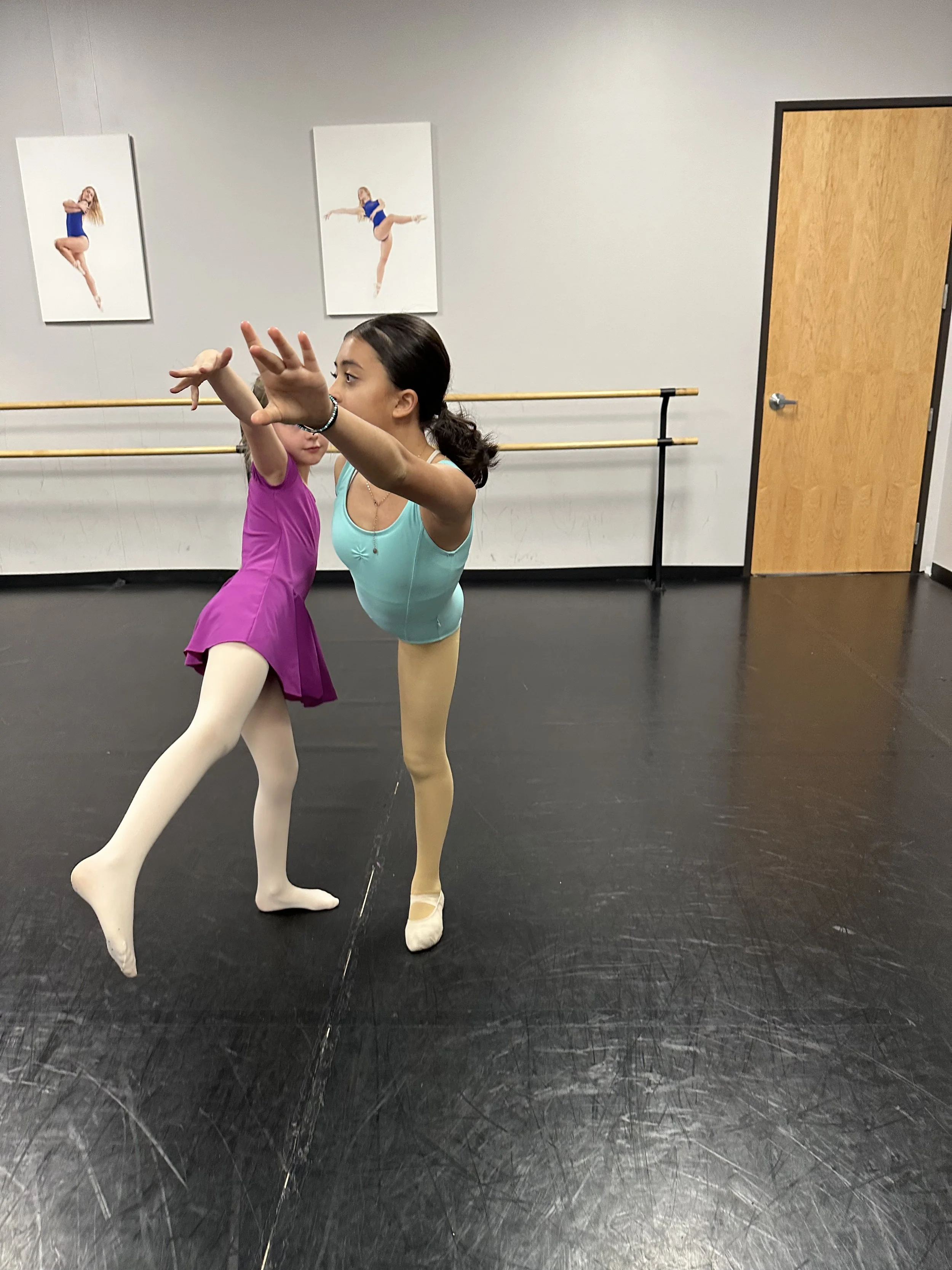 Two young ballet dancers practicing in a dance studio, with posters of dancers on the wall behind them, wooden ballet barres, and a wooden door.
