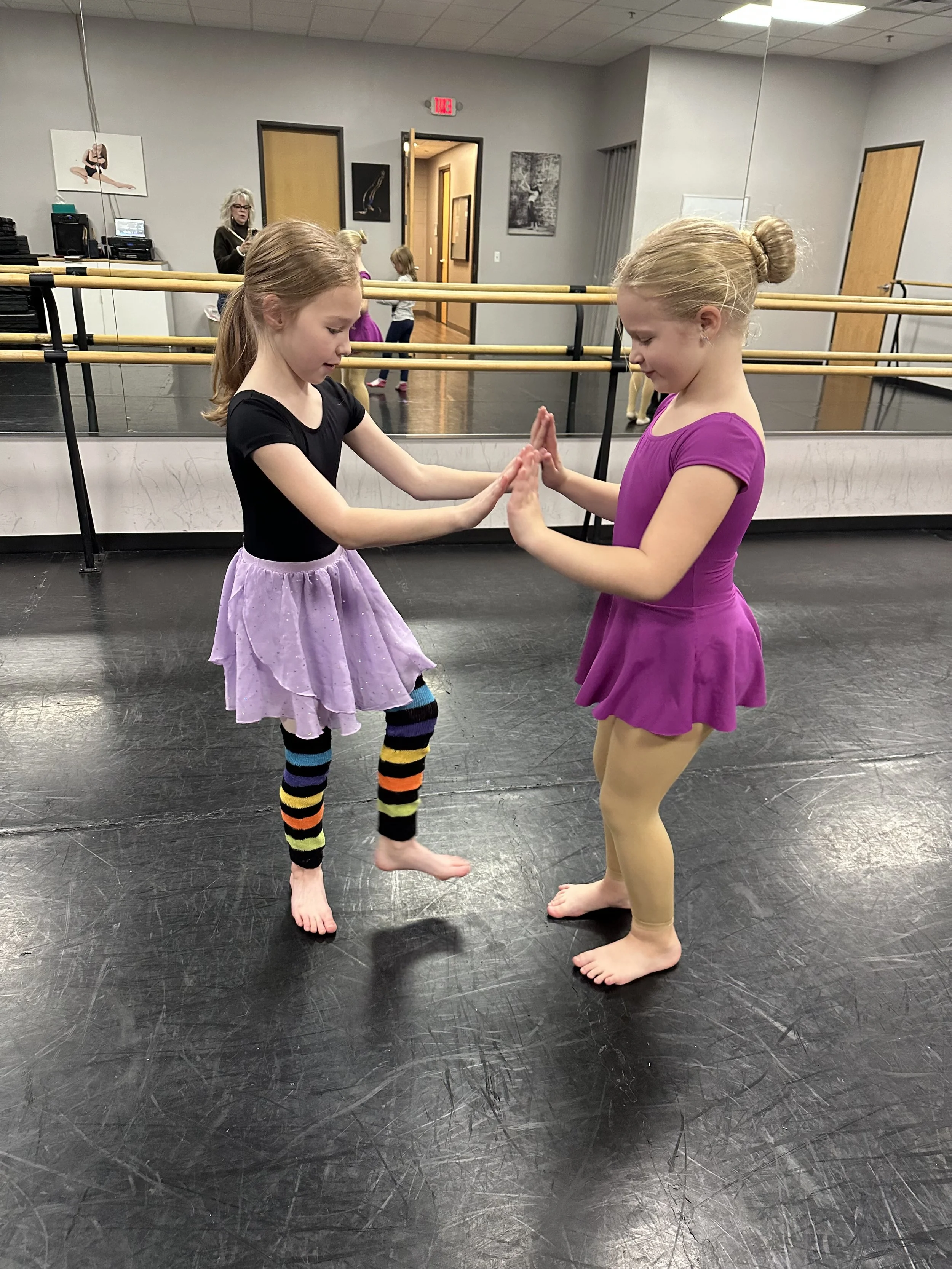 Two young girls are in a dance studio practicing ballet, with one girl in a black shirt and purple skirt and the other in a magenta top and beige leggings. They are standing barefoot facing each other with their palms touching, engaging in a dance ac