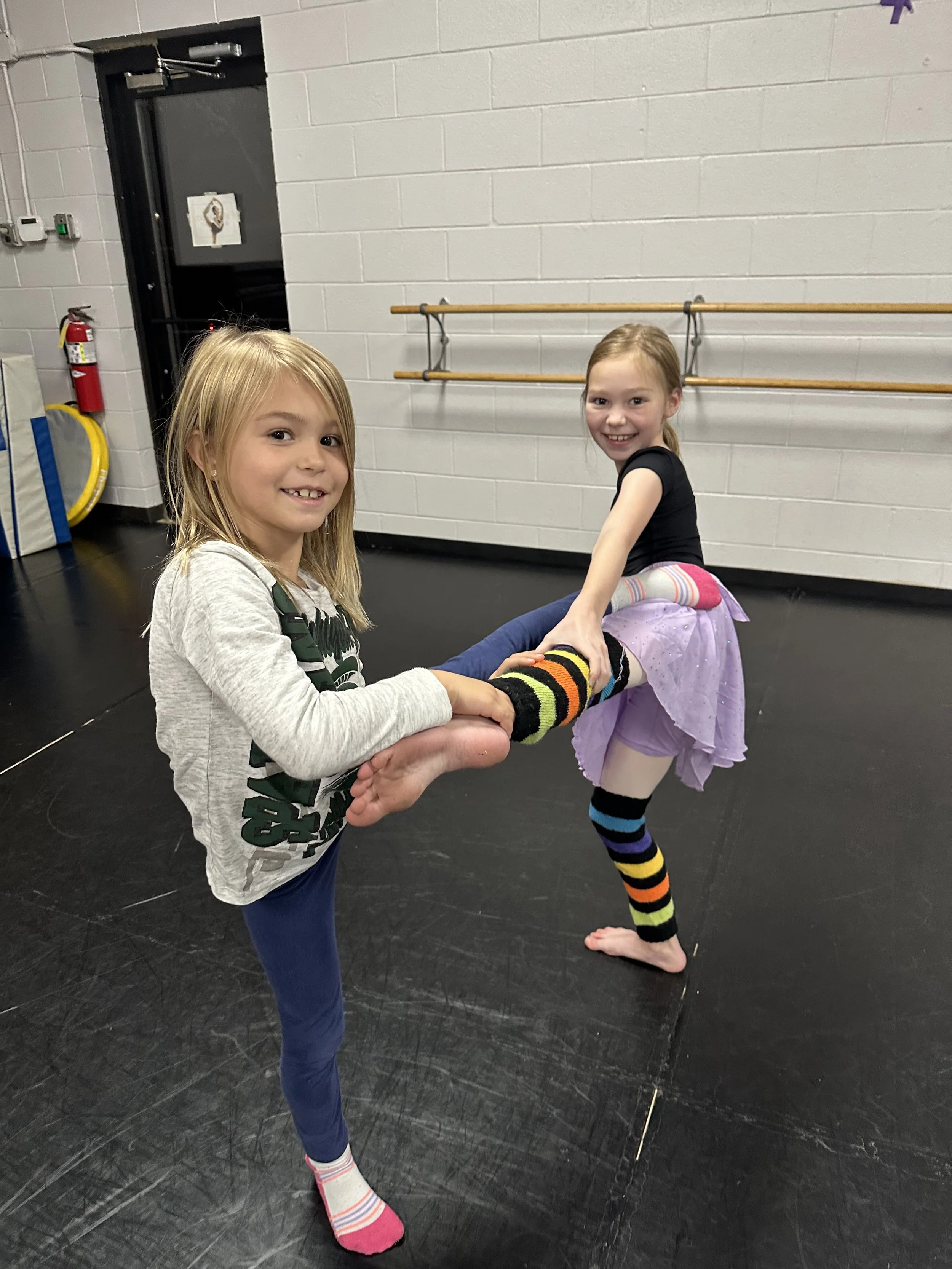 Two young girls holding hands in a dance studio, one with a gray shirt and navy pants, the other with a black shirt and purple tutu, both smiling, with ballet barres and gym equipment in the background.