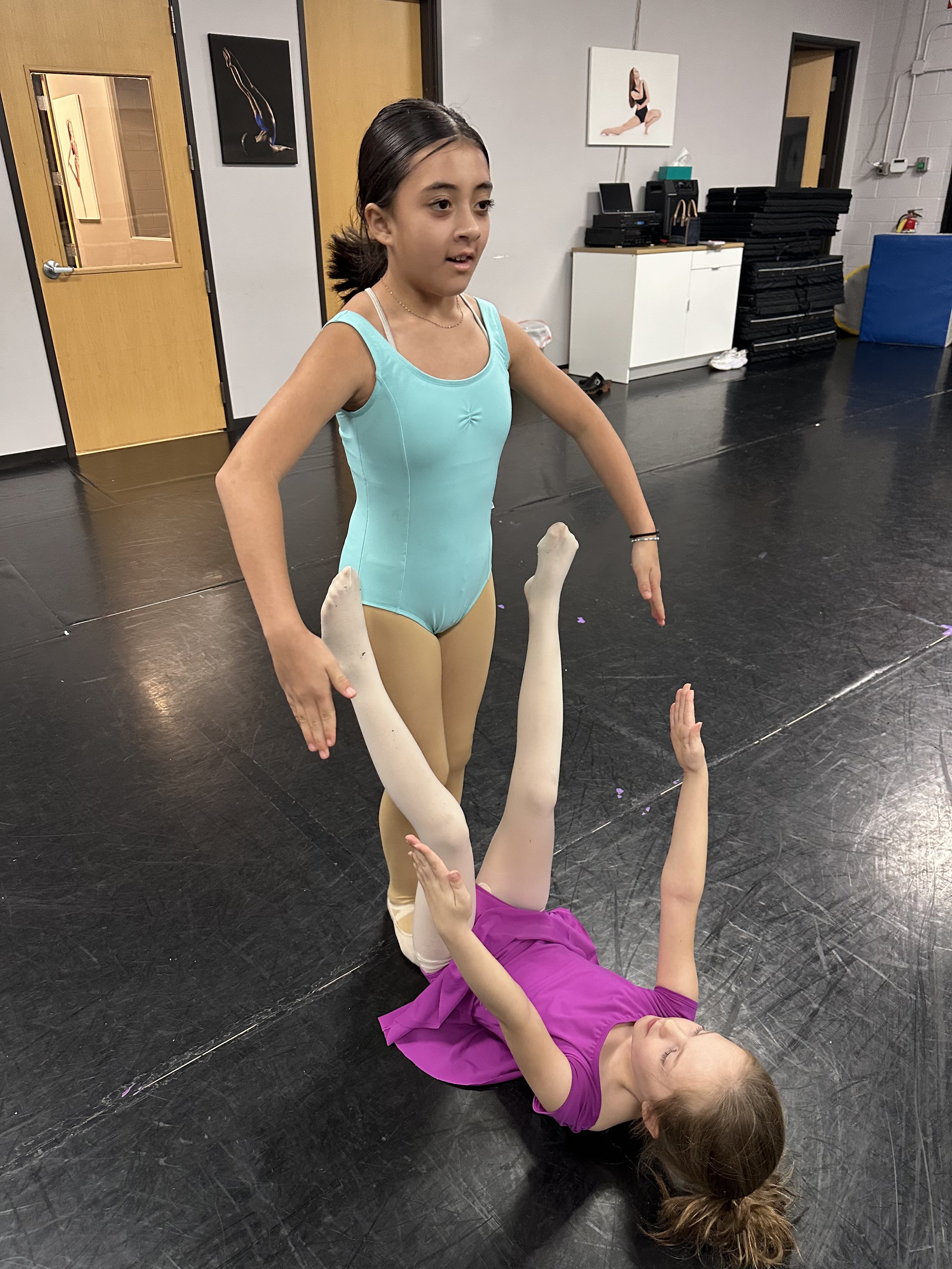 Two young girls in ballet attire practicing ballet in a dance studio. One girl in turquoise leotard standing over another girl in a purple dress lying on the floor. The studio has black flooring, white walls, and ballet posters on the walls.