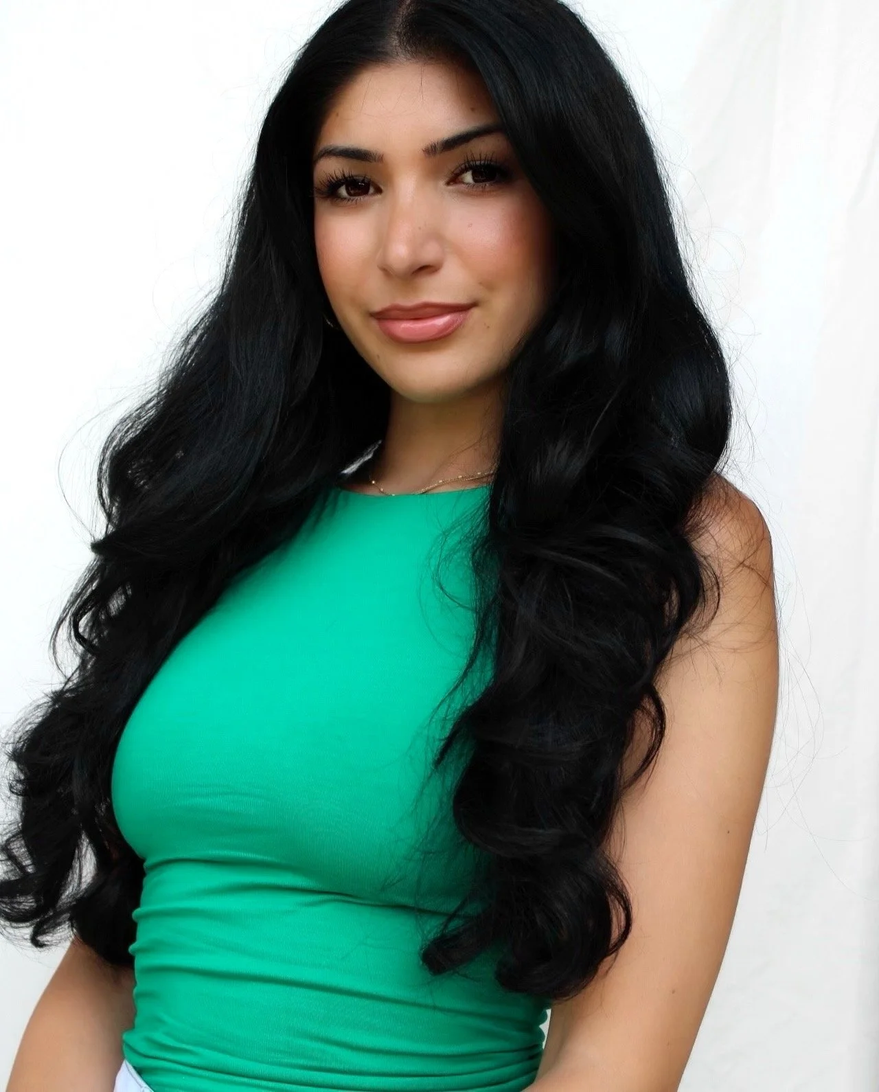 A woman with long, wavy black hair wearing a sleeveless green top standing against a plain white background.