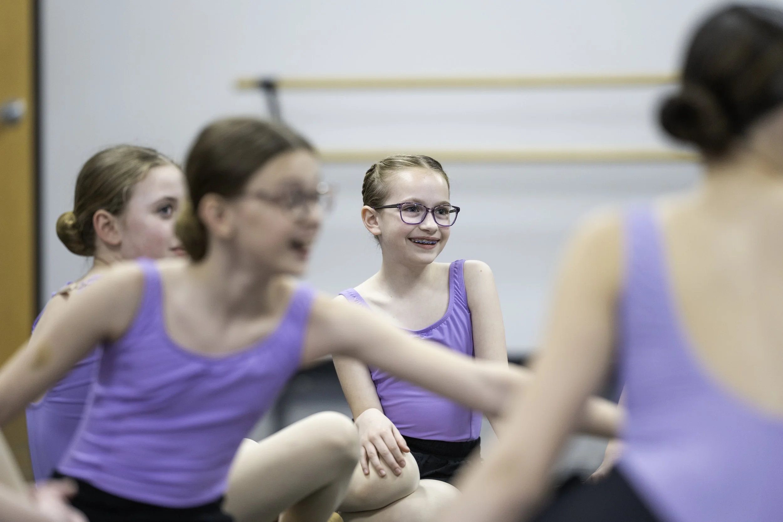 Young girls in purple leotards sitting on the floor in a dance class, smiling and talking to each other.