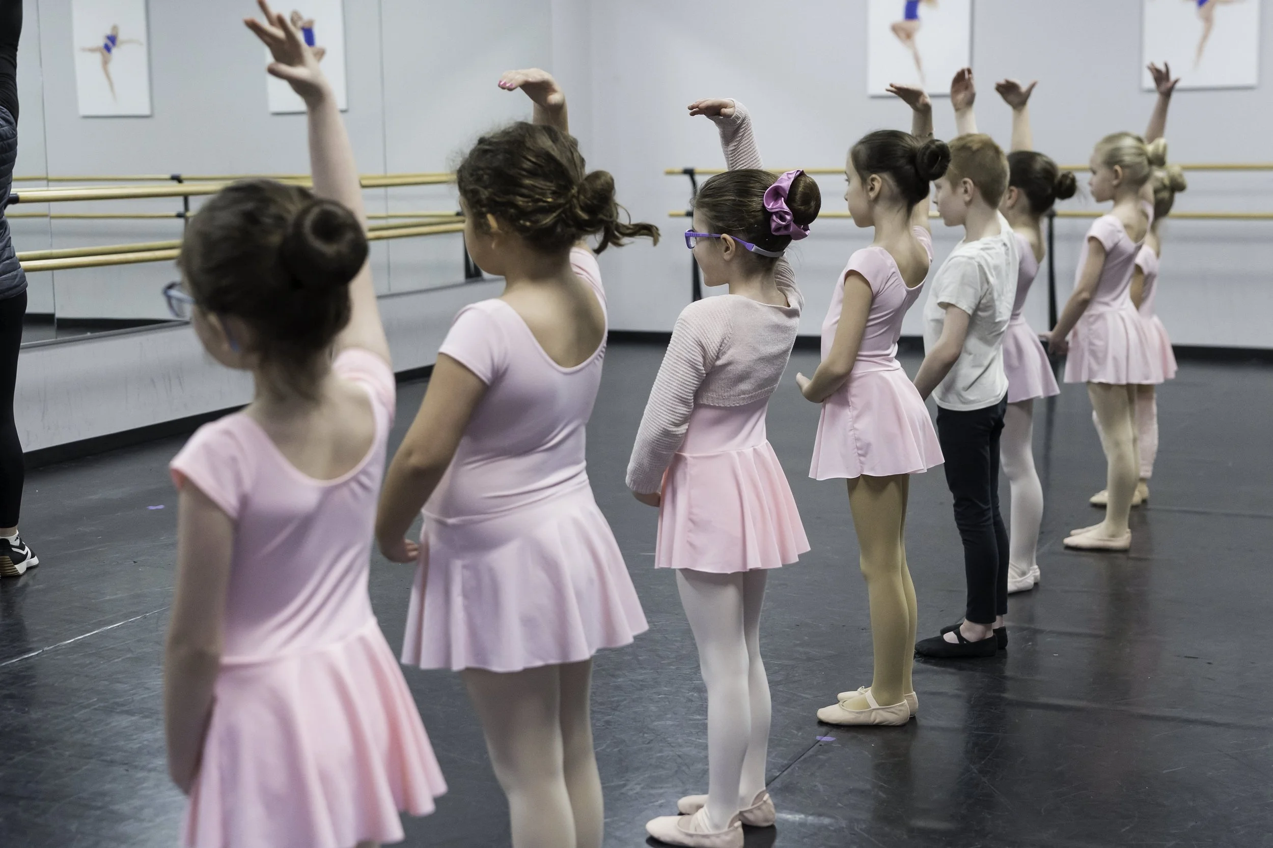 Young ballet students in pink costumes and ballet slippers stand in a line at a dance studio, practicing ballet moves with their arms raised. The studio has mirrors and ballet barres.