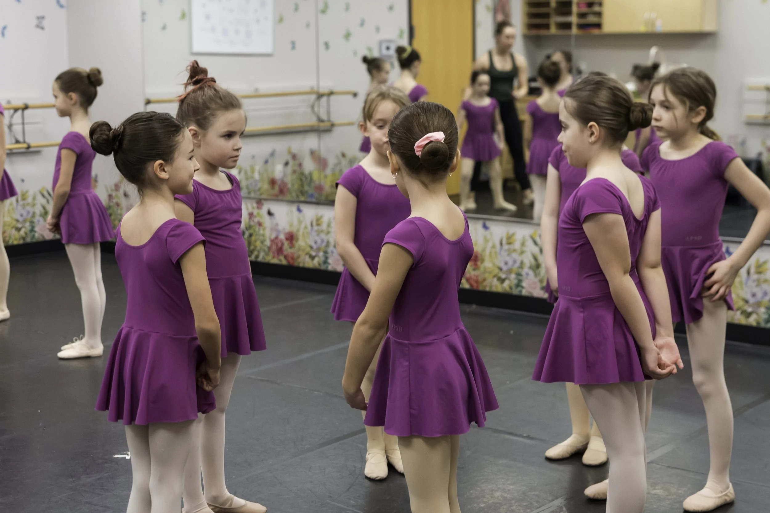 Young girls in purple ballet dresses practicing in a dance studio with mirrors and ballet barres.