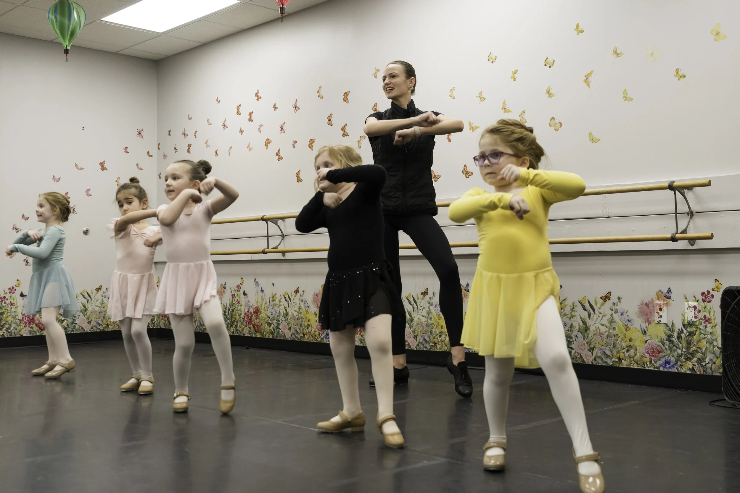 A ballet class with six young girls in colorful leotards and an instructor practicing dance moves in a studio decorated with butterfly and floral wall art.