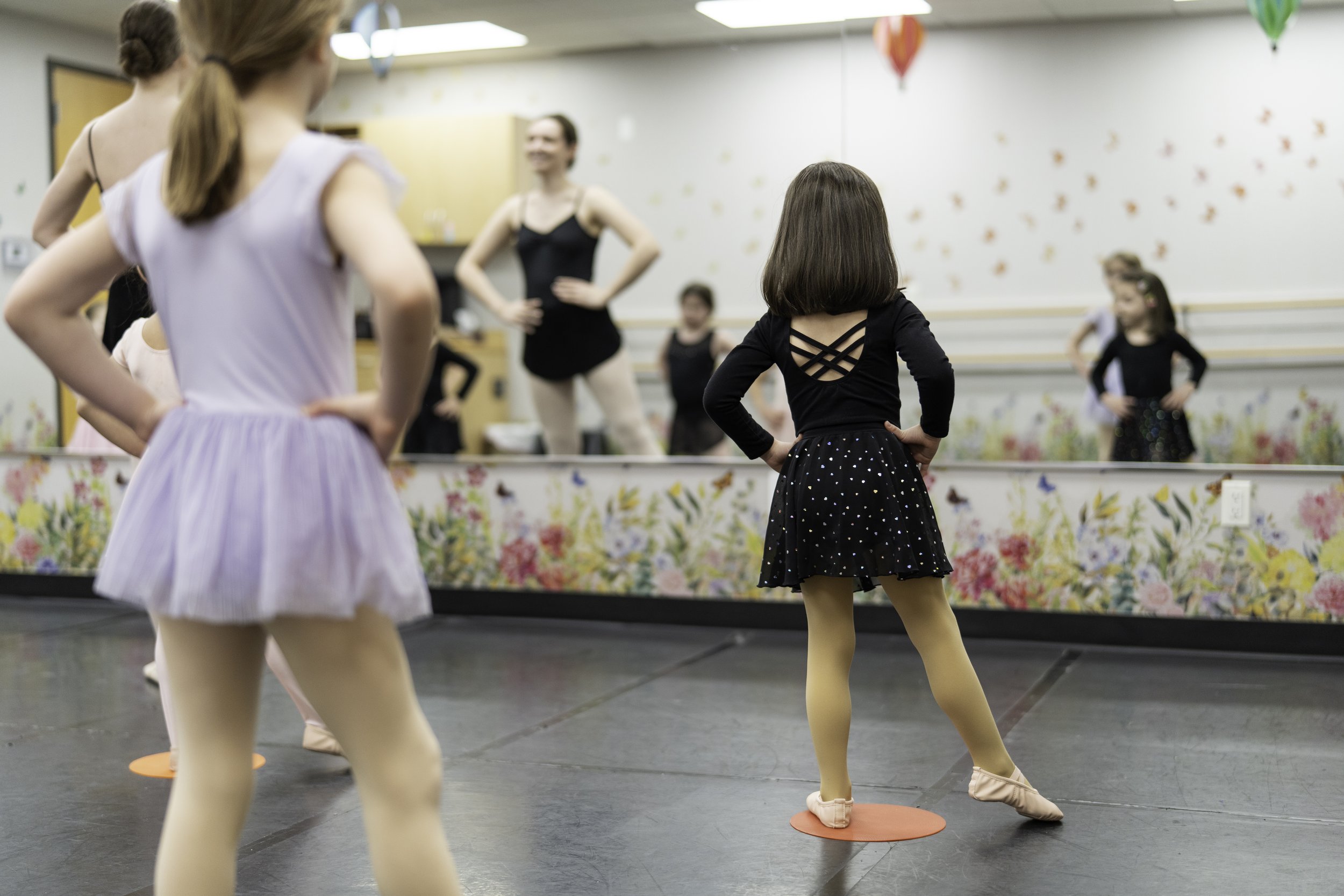 Ballet class with young girls practicing in front of a mirror, wearing costumes and ballet slippers, with an instructor guiding.
