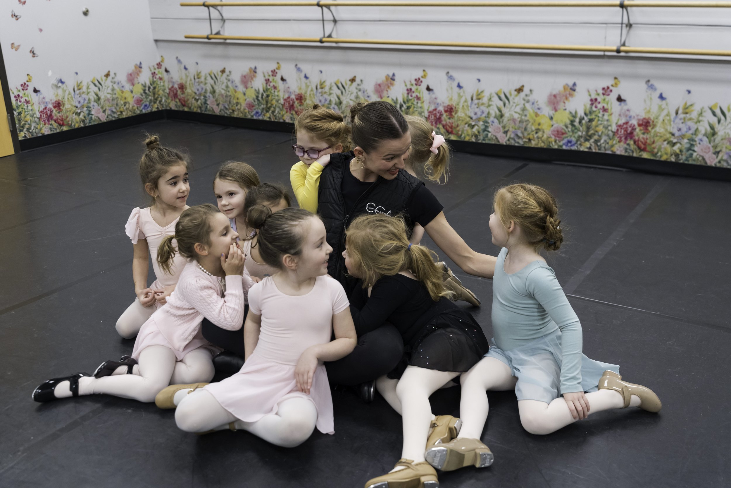 A group of young girls in ballet costumes sitting and kneeling on the floor, gathered around their ballet instructor in a dance studio with floral wall decor.