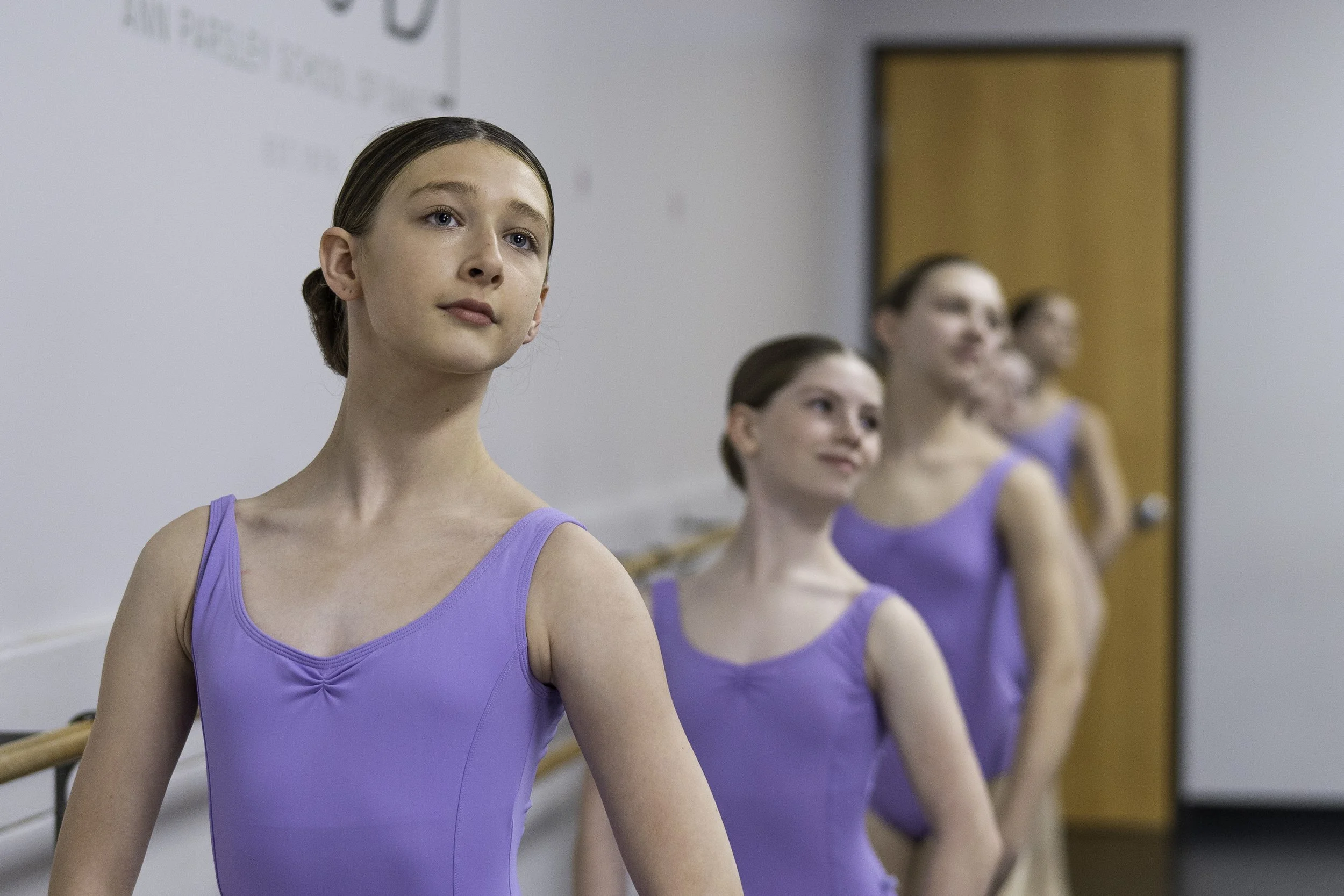 Four young girls in purple ballet leotards standing in a line at a ballet studio, facing the same direction. The background includes a white wall with some writing and a wooden door.