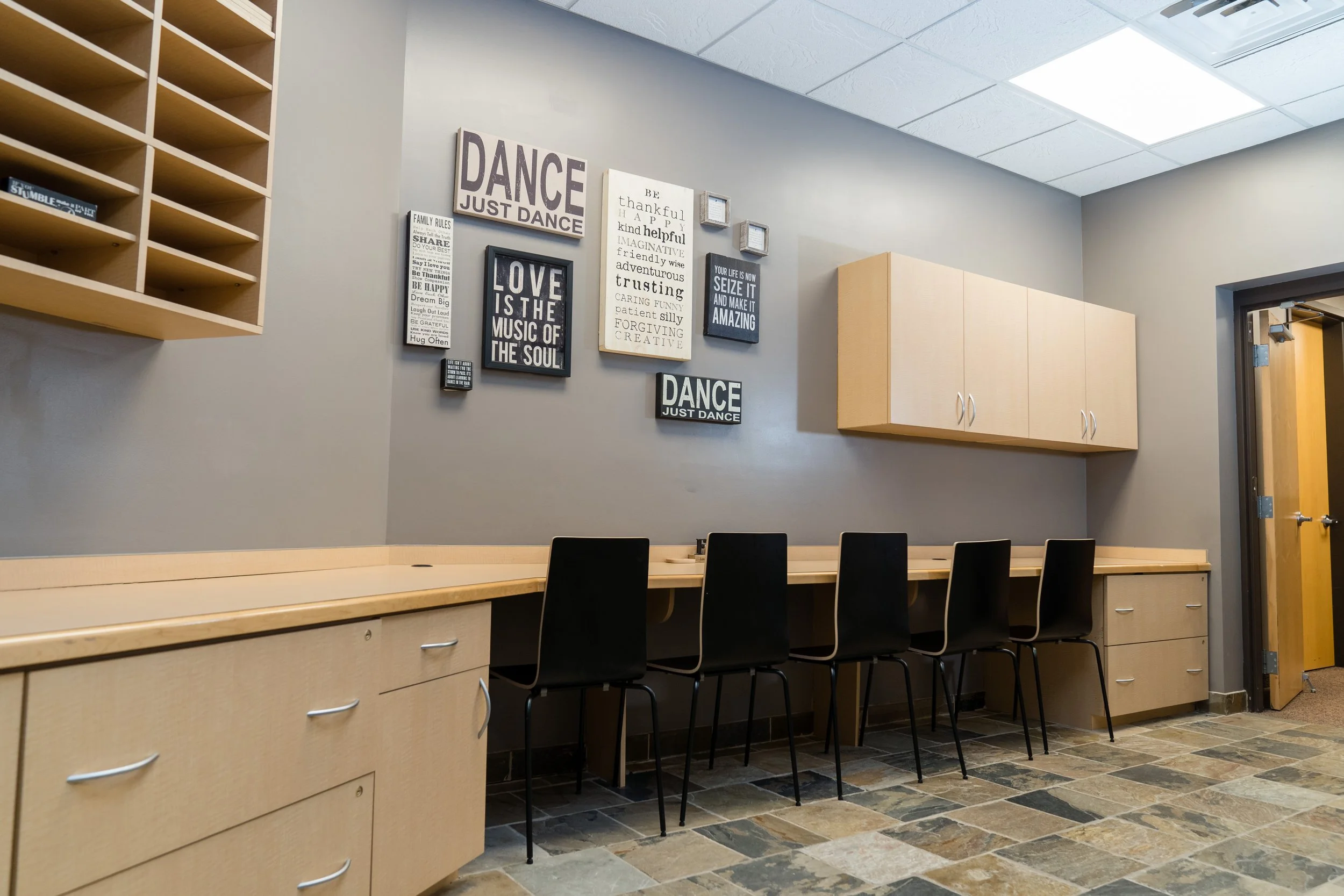 Room with a long wooden counter and five black chairs, decorated with framed motivational and dance-themed posters on a gray wall, with wooden cabinets and a wooden door.