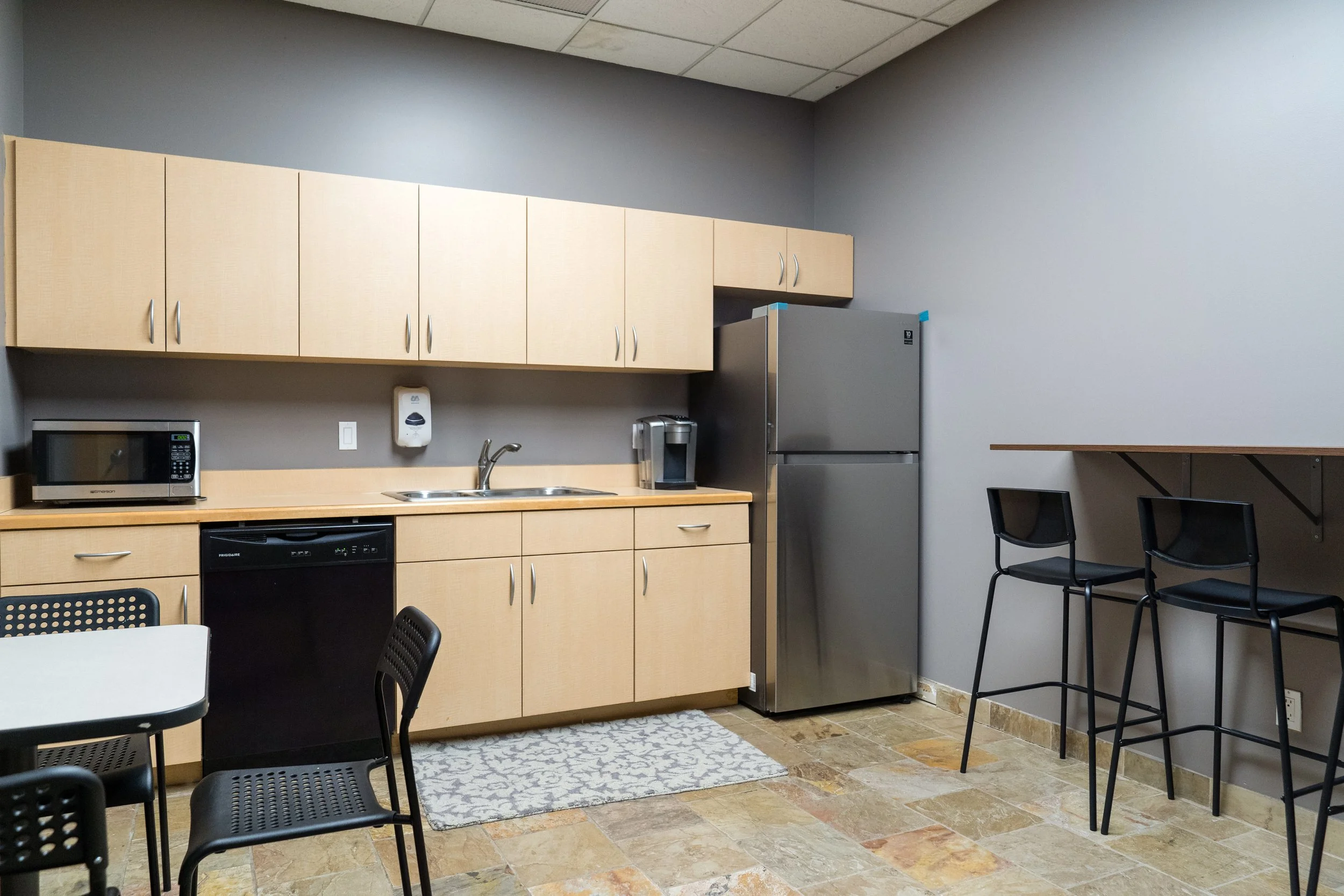 A kitchenette with beige cabinets, a refrigerator, microwave, coffee maker, black dishwasher, sink, and small tables with black chairs in a room with tiled floor and gray walls.