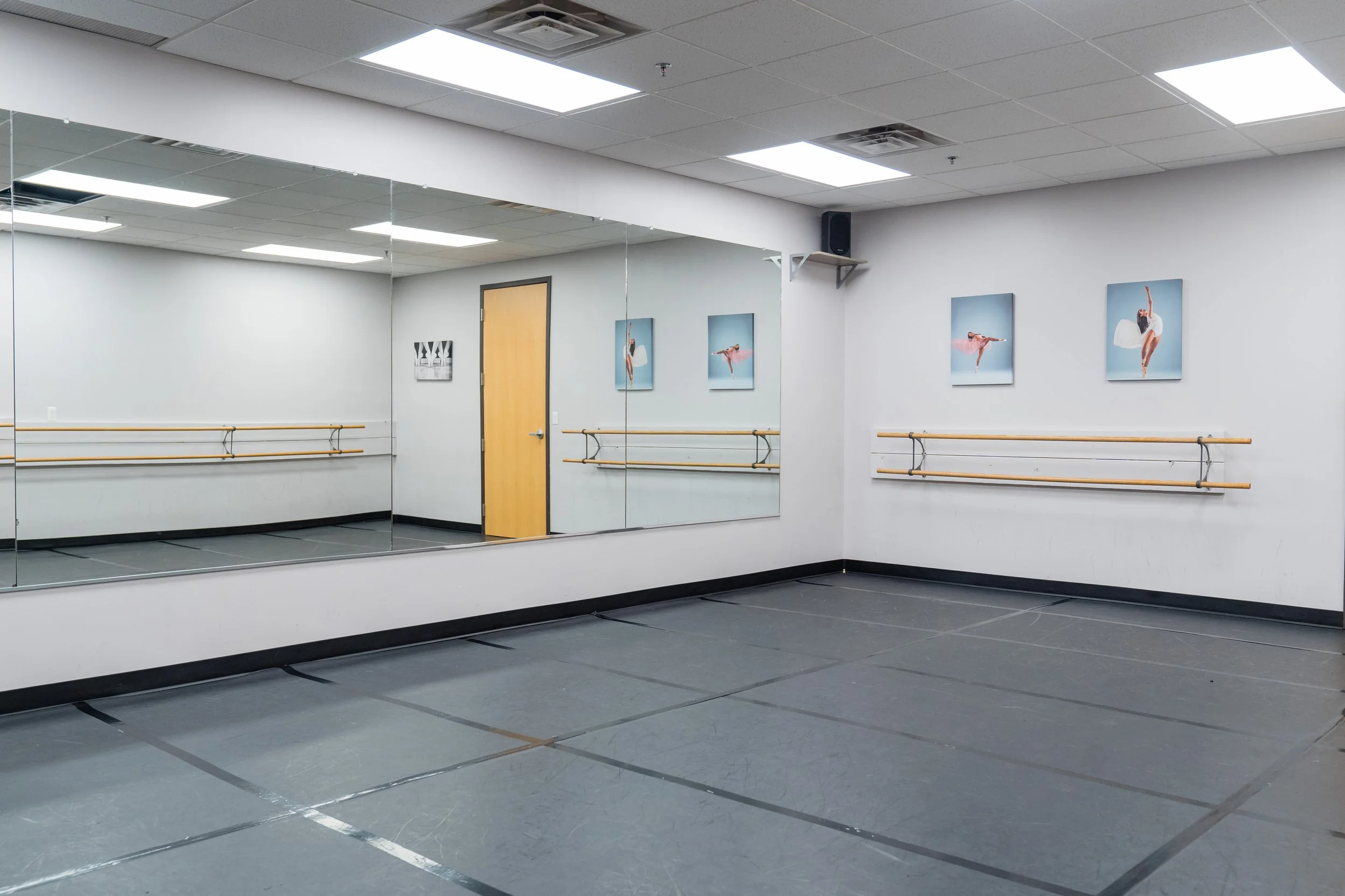 Dance studio with mirrored wall, ballet barres, framed ballet pictures, wooden door, and black flooring.