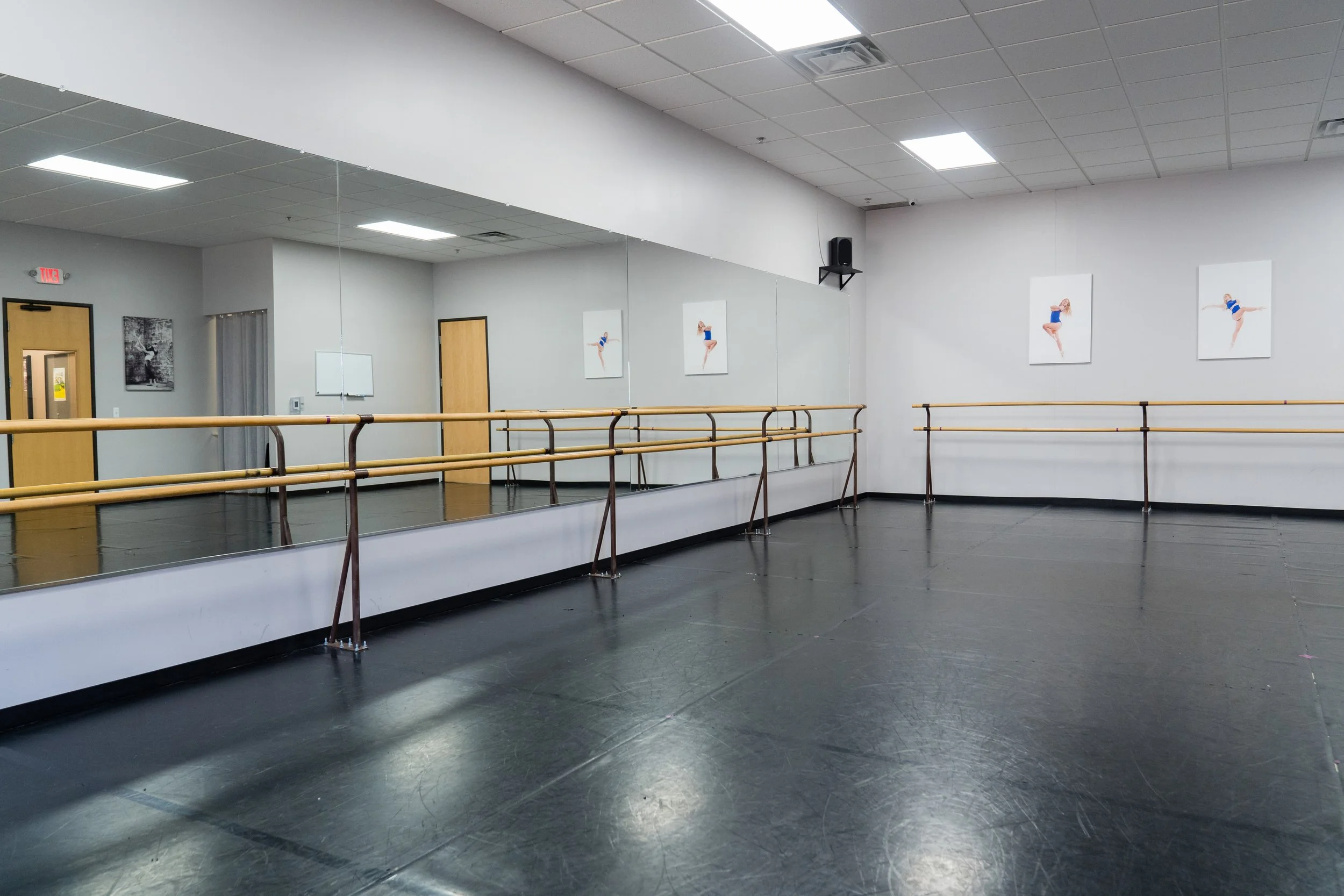 Empty dance studio with black flooring, wooden ballet barres, and large mirrors on one wall. Four posters of dancers in blue costumes are on the walls.