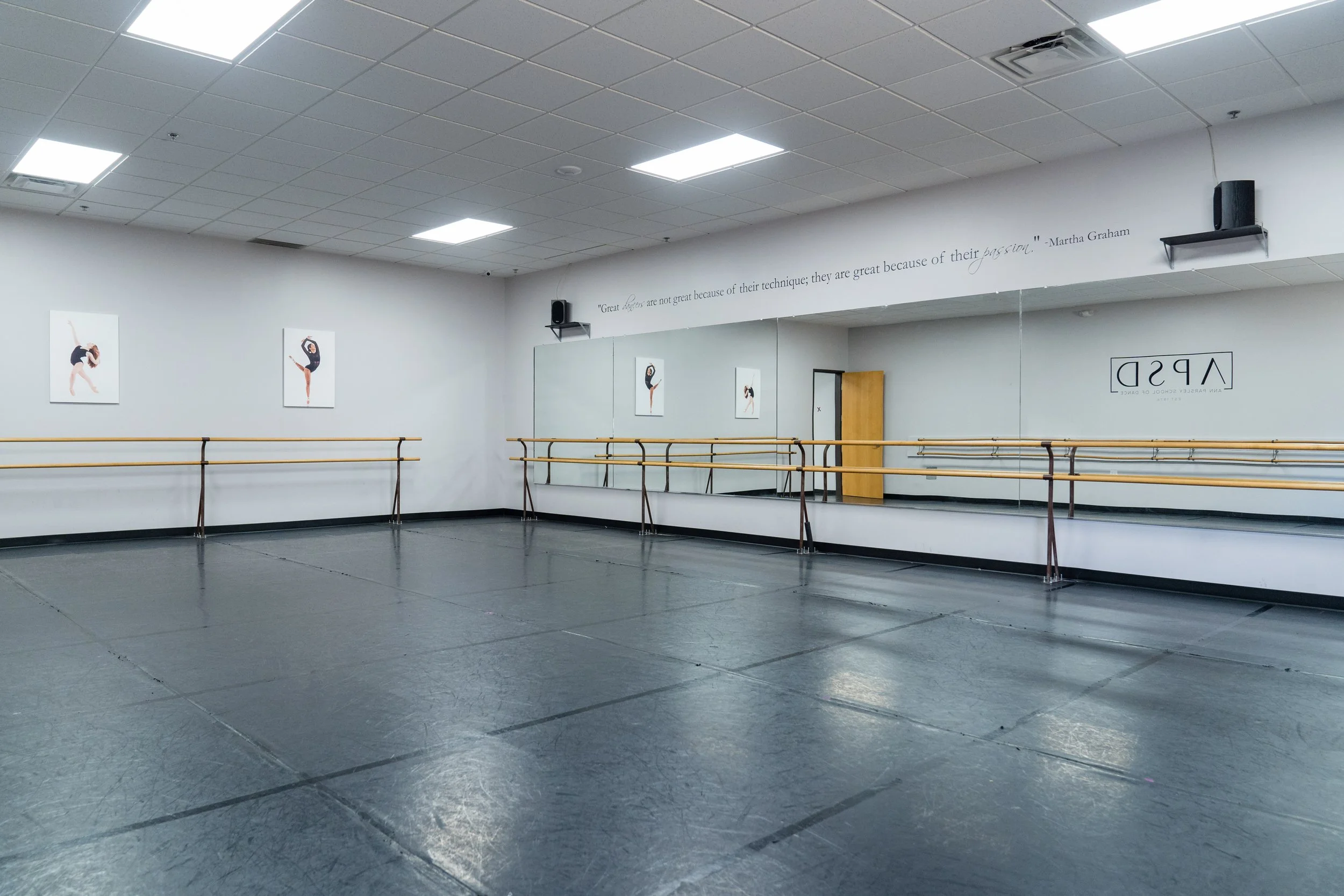 Empty dance studio with black floor, white walls, ballet barres, large mirror, and three dance posters