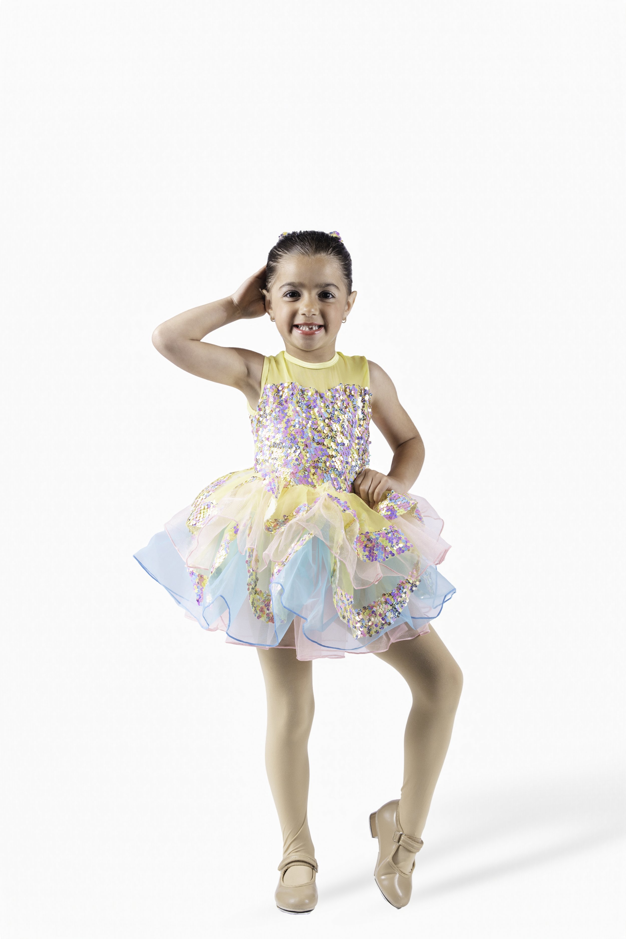Young girl in a colorful, glittery dress with a tutu, beige dance shoes, smiling and posing with one hand behind her head on a white background.