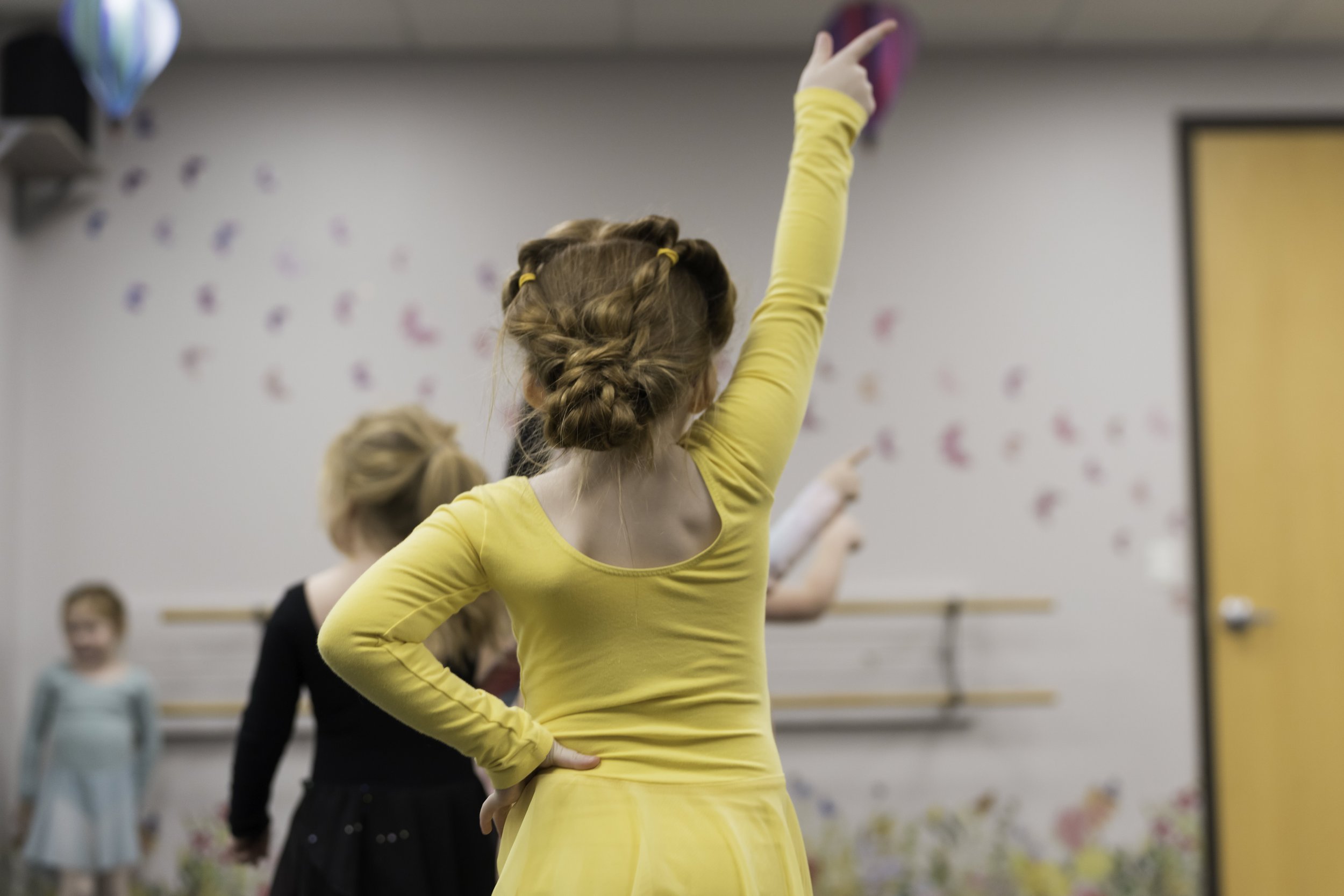 A young girl with red hair styled in braids wearing a yellow dress raises her hand in a dance studio, with other girls in dance costumes in the background.