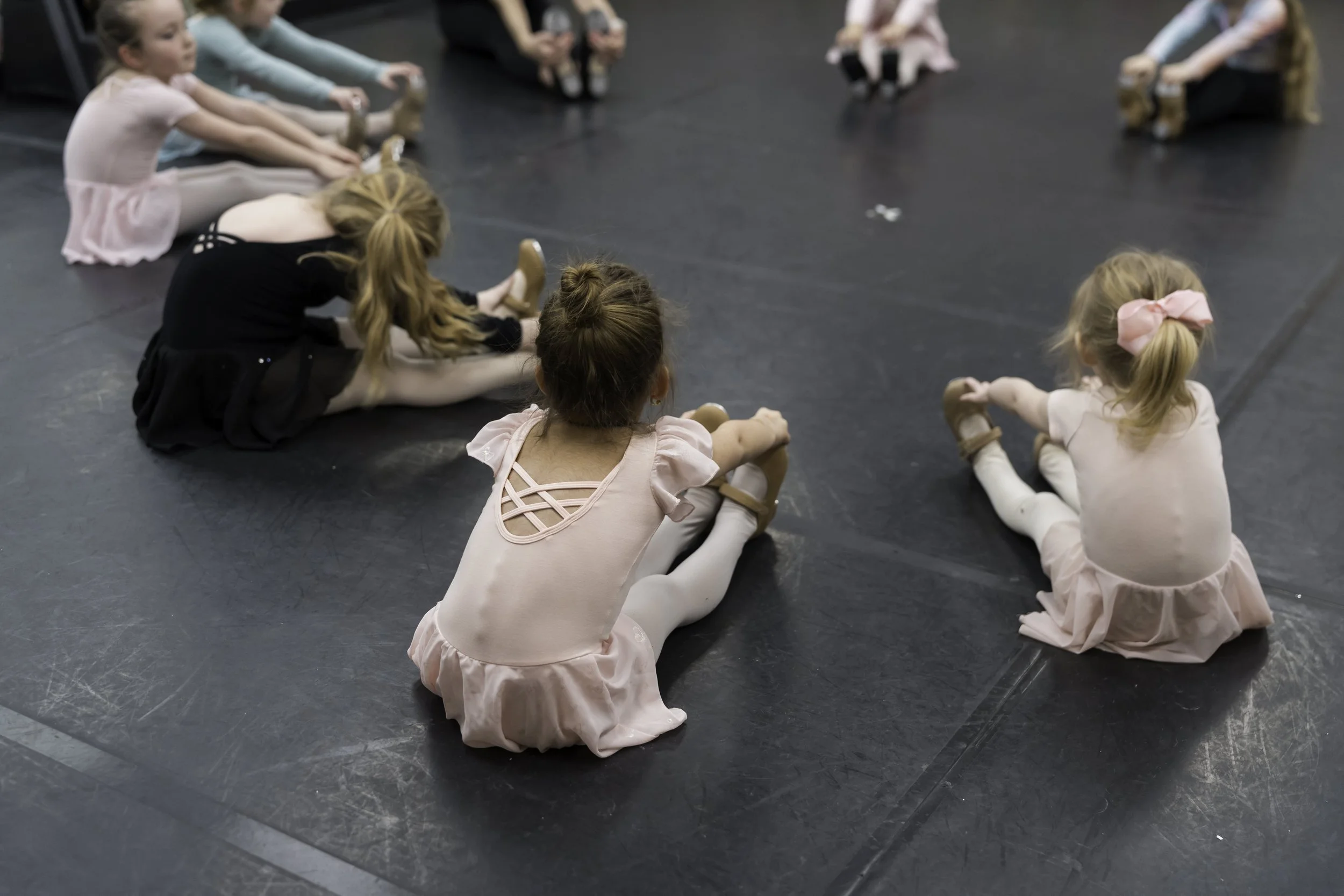 Group of young girls in ballet costumes sitting on a black dance studio floor, stretching in a circle.