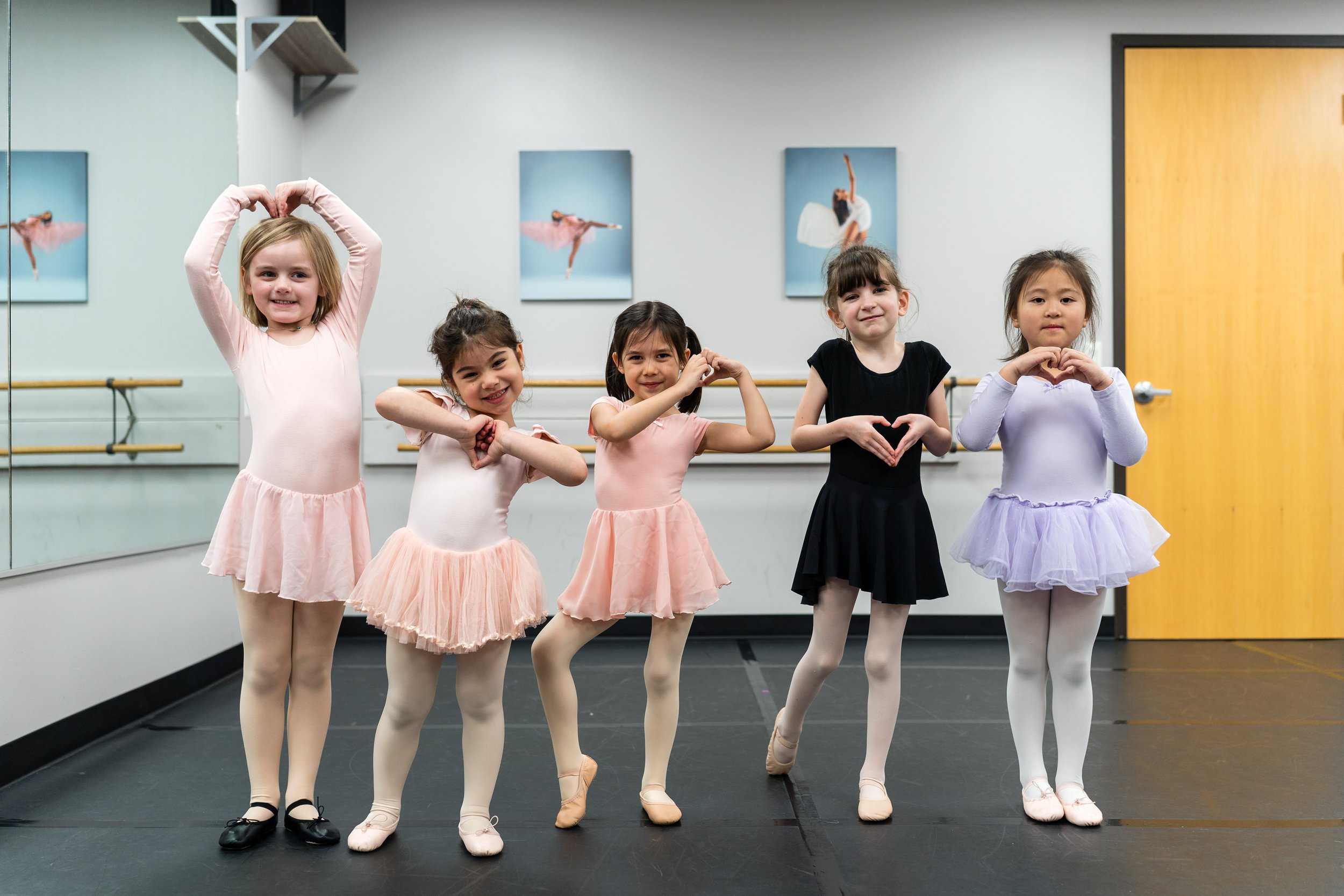 Five young girls in ballet outfits standing in a dance studio, each making a heart shape with their hands and smiling.