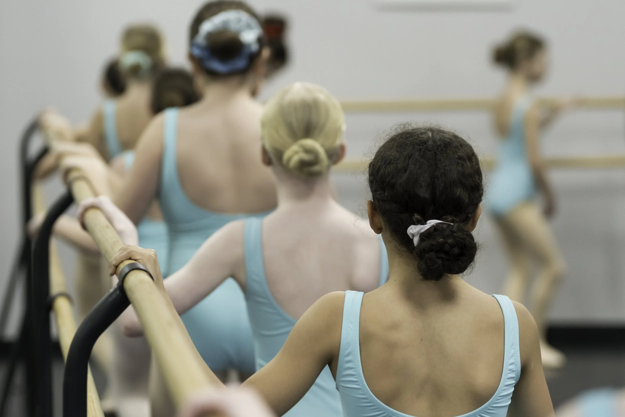 Ballet students in a dance class, wearing blue leotards, with some standing at the barre and one practicing at the mirror in the background.