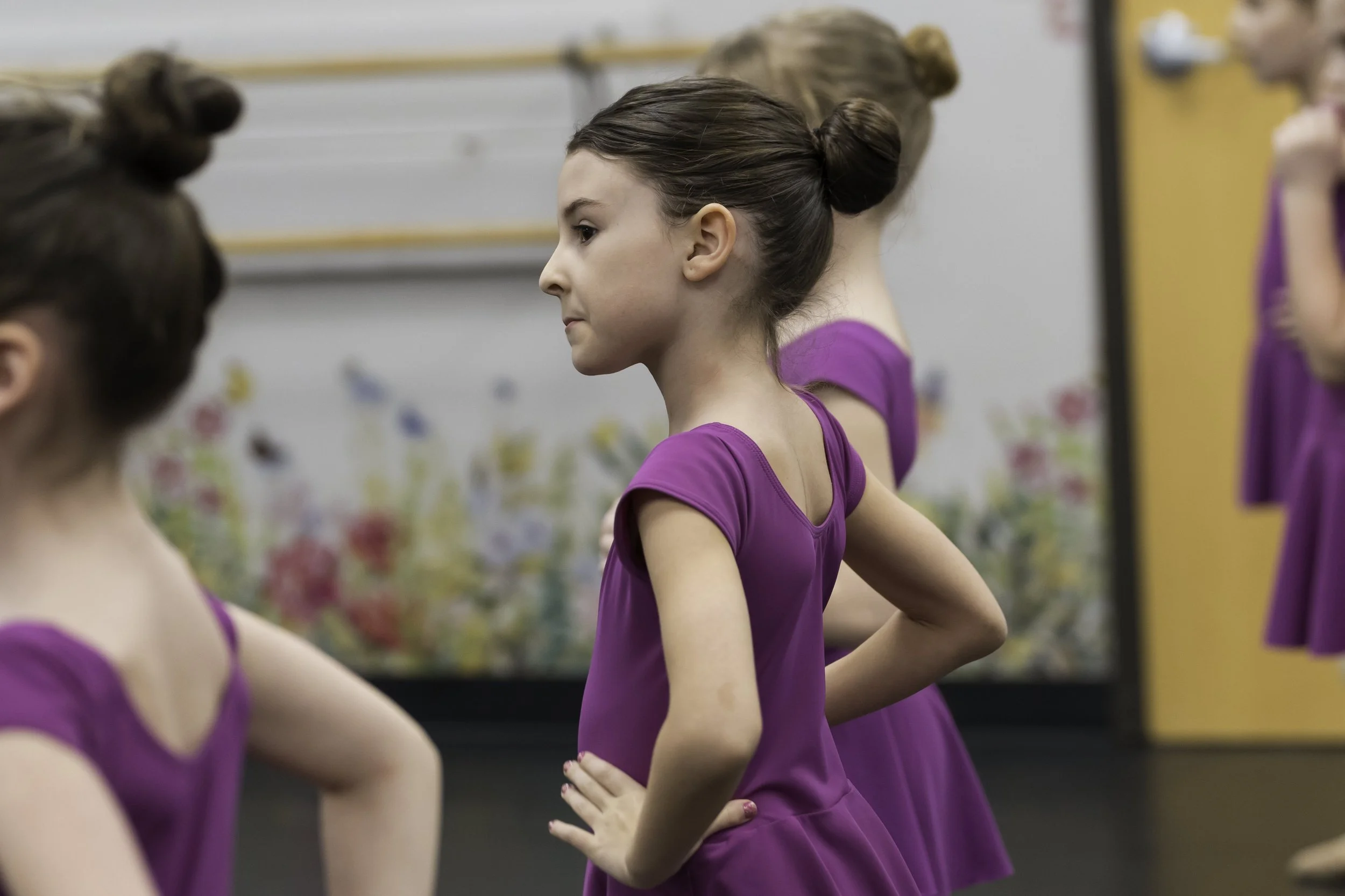 A young girl in a purple dance costume standing with her hands on her hips among other dancers, in a dance studio or classroom with a floral background.