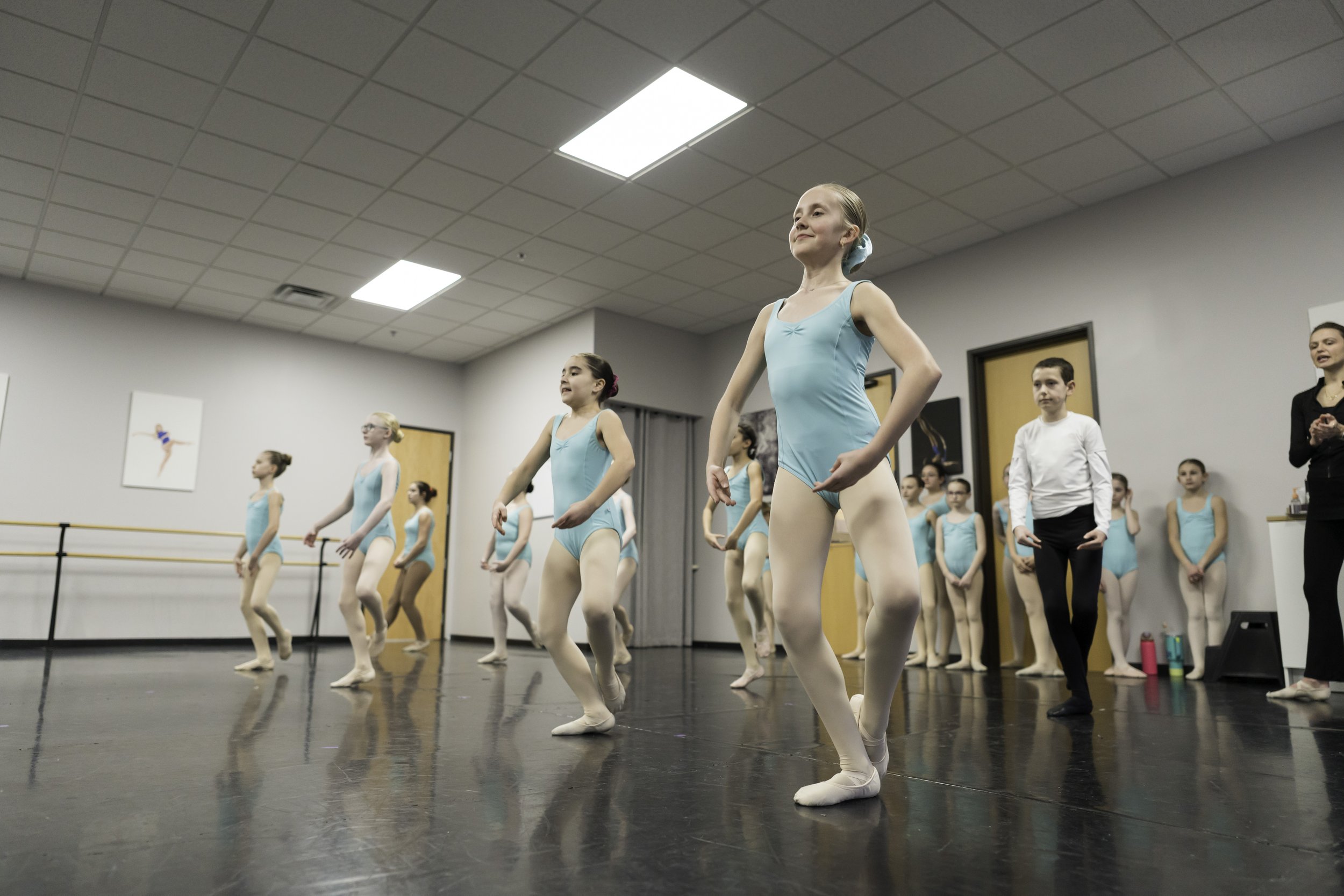 Group of young ballet dancers in blue leotards and white tights practicing in a dance studio, with their instructor observing.