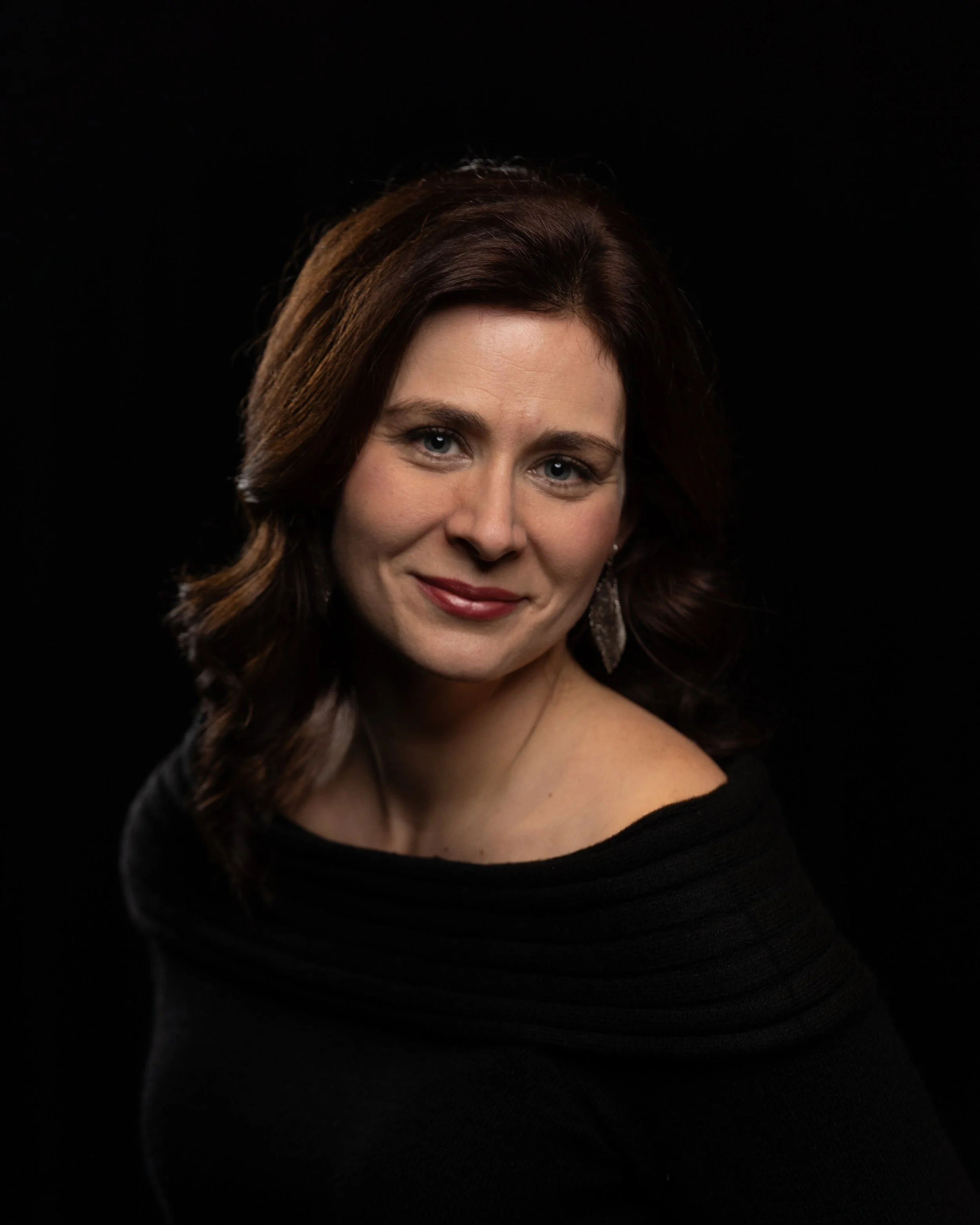 Portrait of a woman with shoulder-length brown hair wearing a black top and silver earrings, smiling against a black background.