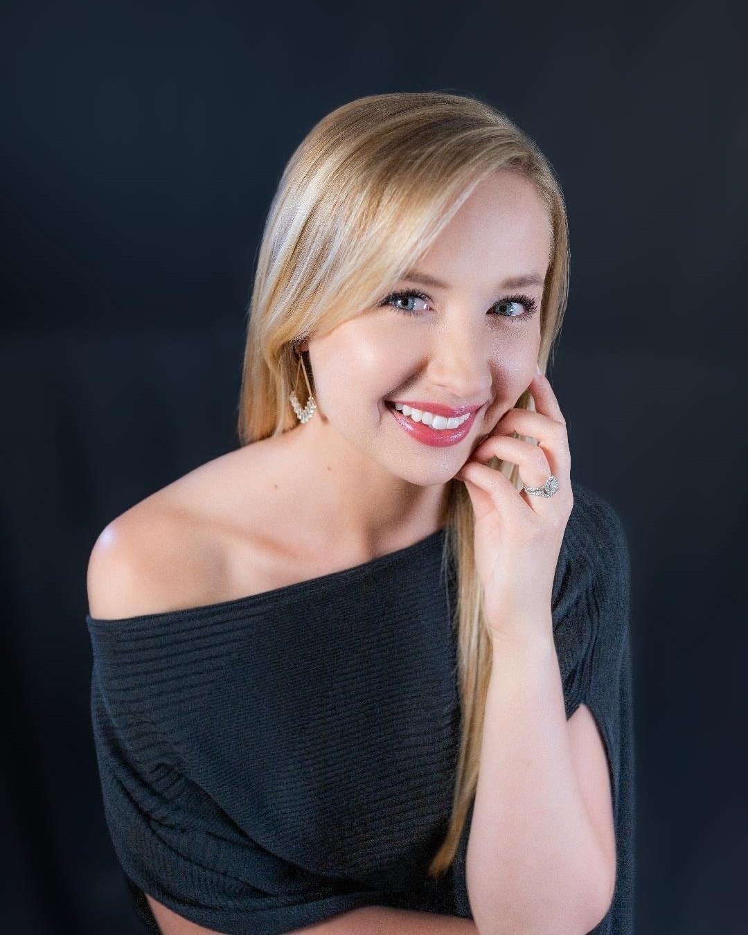 A smiling woman with blonde hair wearing a black off-the-shoulder top, silver earrings, and a ring, posing against a dark background.